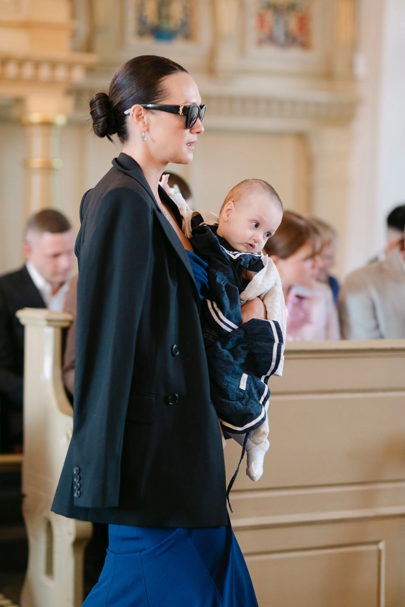 En kvinna med mörkt hår i en knut, mörka solglasögon och smycken, bär ett spädbarn under en ceremoni i en kyrka.  Av Sofia Wilhelmina bröllopsfotograf i Stockholm.