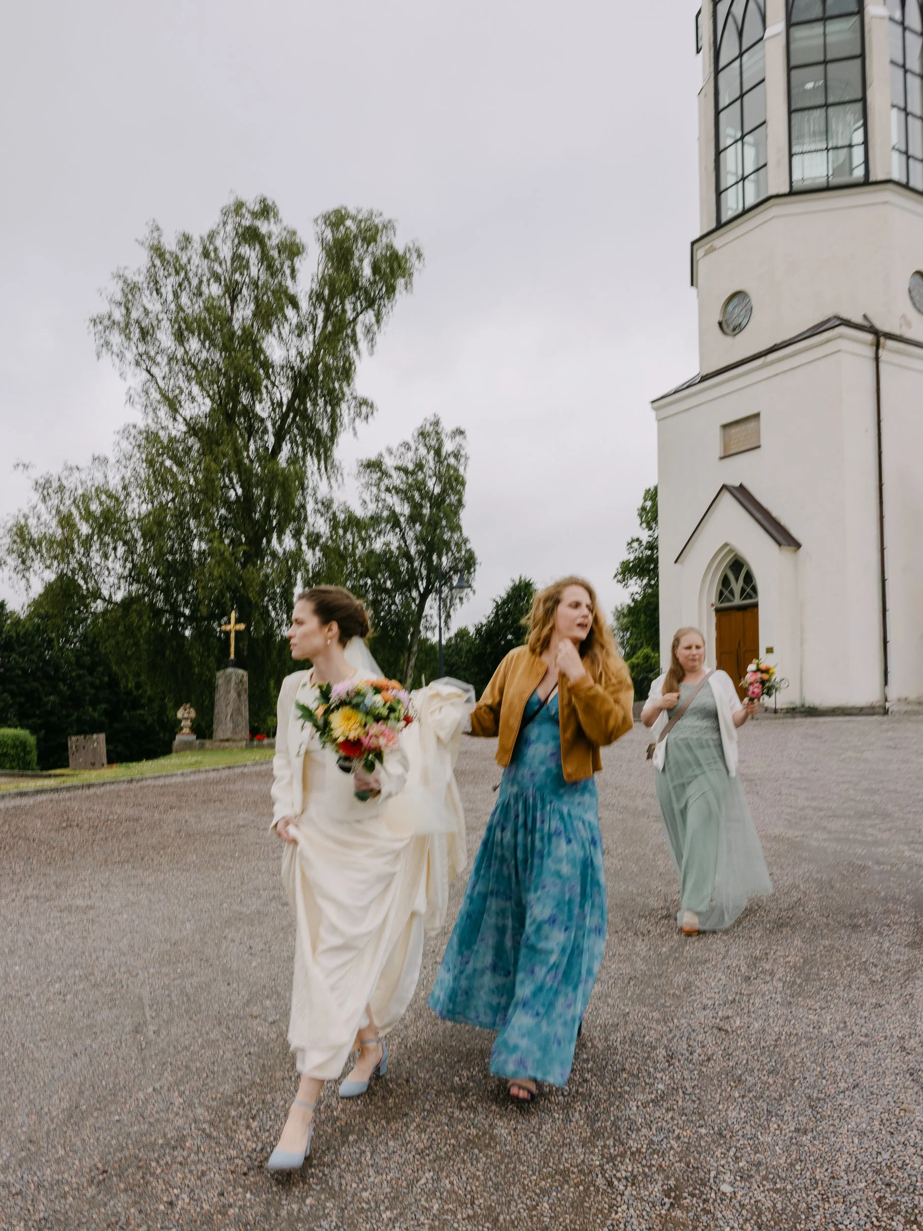 Tre kvinnor i bröllopskläder som går utanför en vit kyrka med gröna träd i bakgrunden, håller i buketter av blommor, en av kvinnorna bär en vit klänning och en annan en blå klänning.