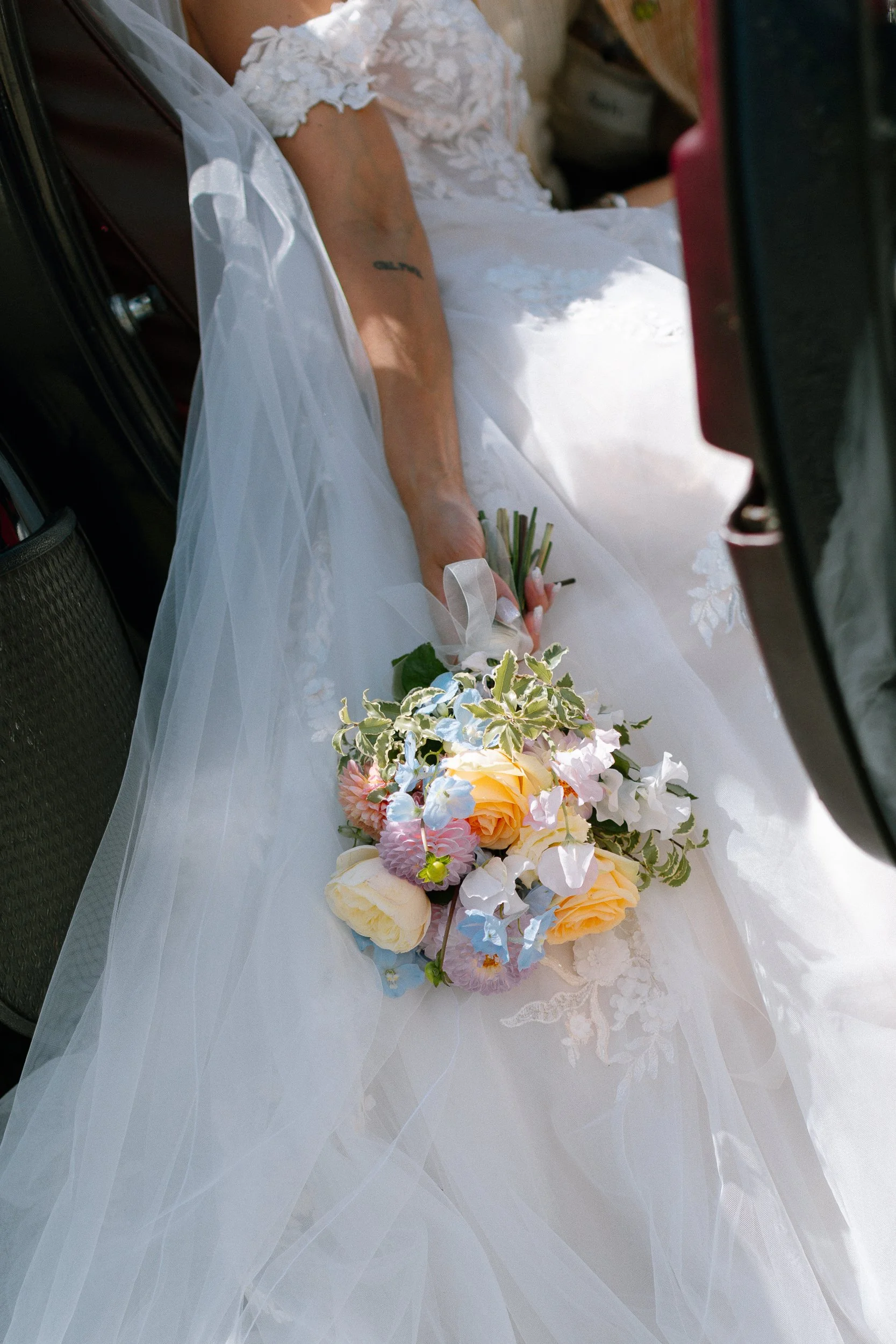 Brudens arm med ett broderat armband och en bukett med ljusa blommor, liggande på ett vitt bröllopsklänningskjol i en bil.