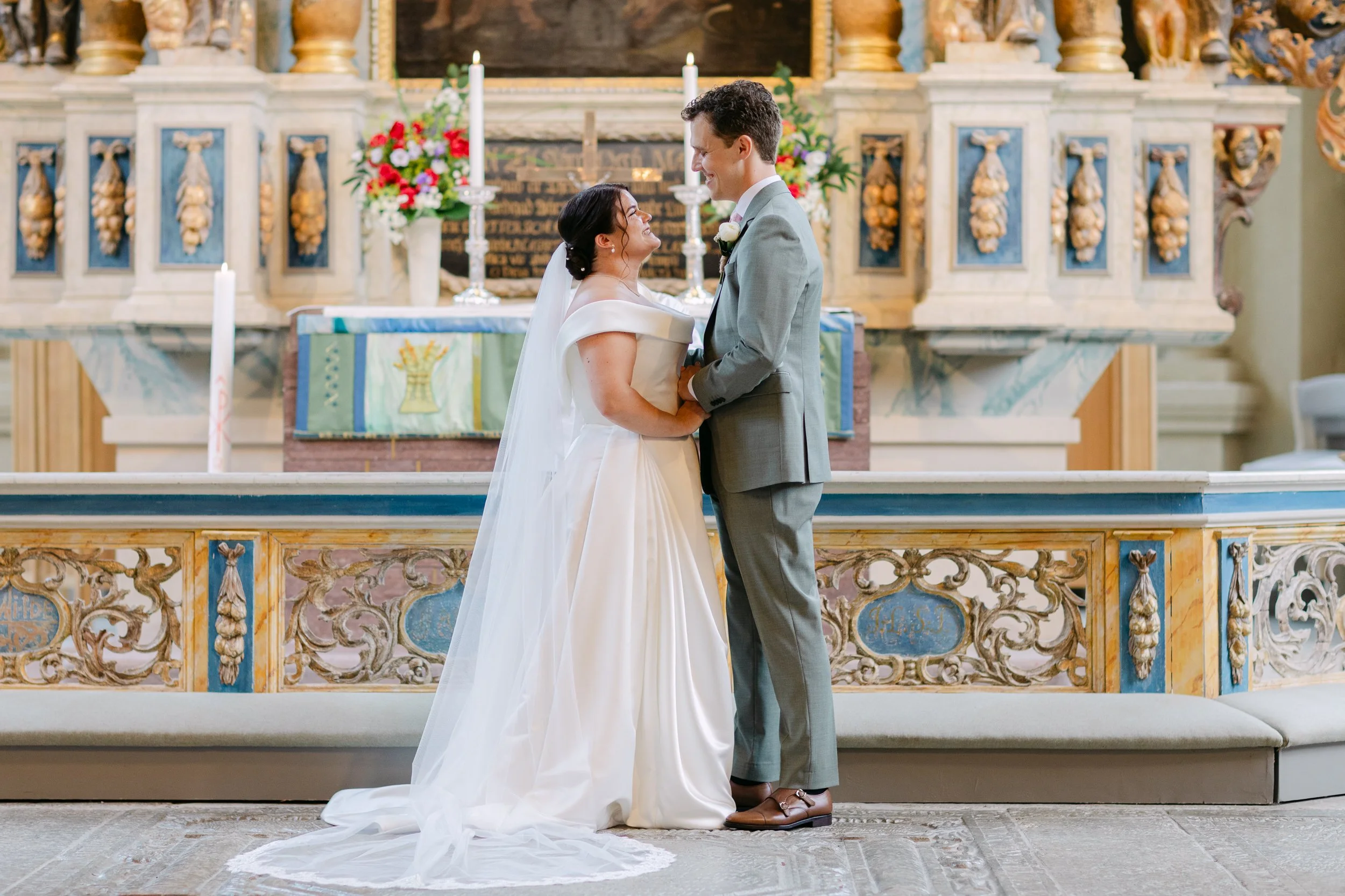 En brud och en man som håller varandra i händerna i en kyrka under ett bröllopsceremoni, omgivna av blomsterarrangemang och ett altare.