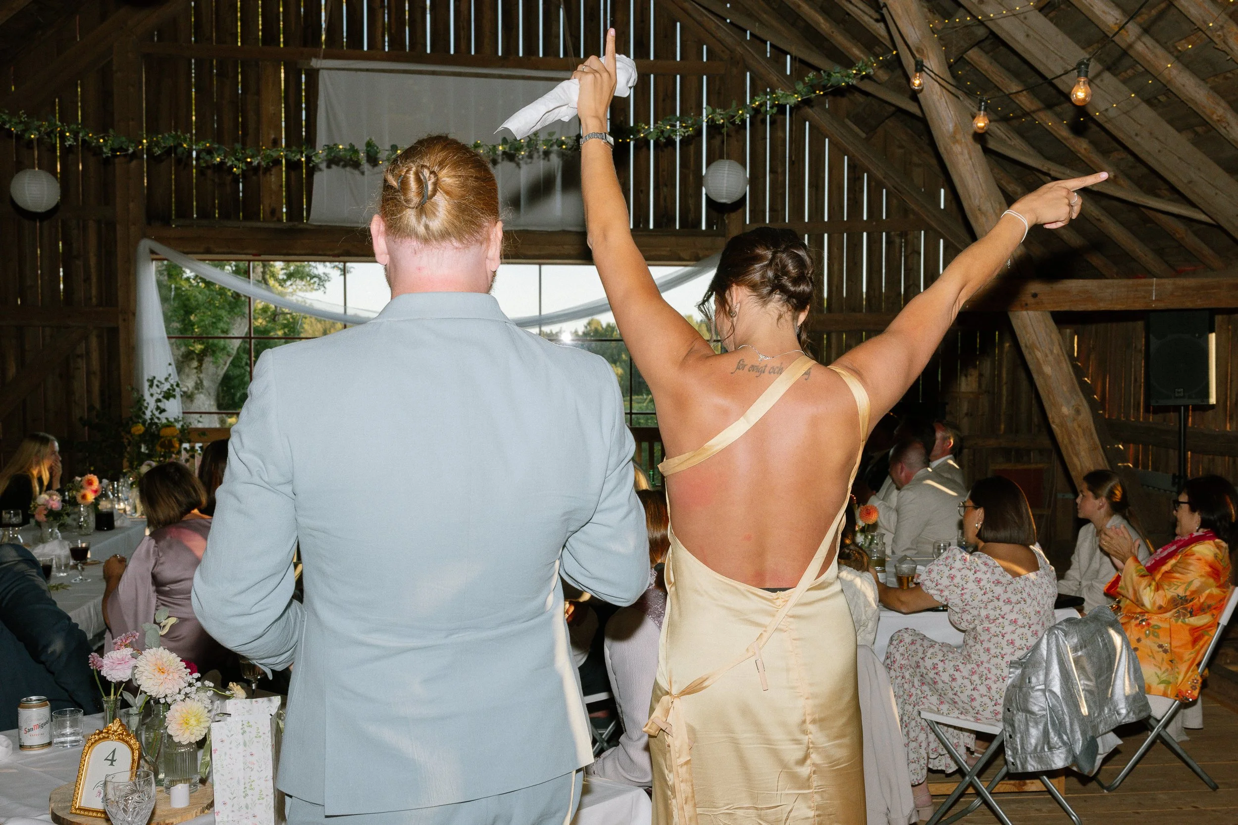 Två personer, en man i ljusblå kostym och en kvinna i en gul klänning, dansar på en fest på en loge. Kvinnan har armarna utsträckta och ser ut att sprudla av glädje, medan gäster sitter vid bord runt dem. Av Sofia Wilhelmina bröllopsfotograf 