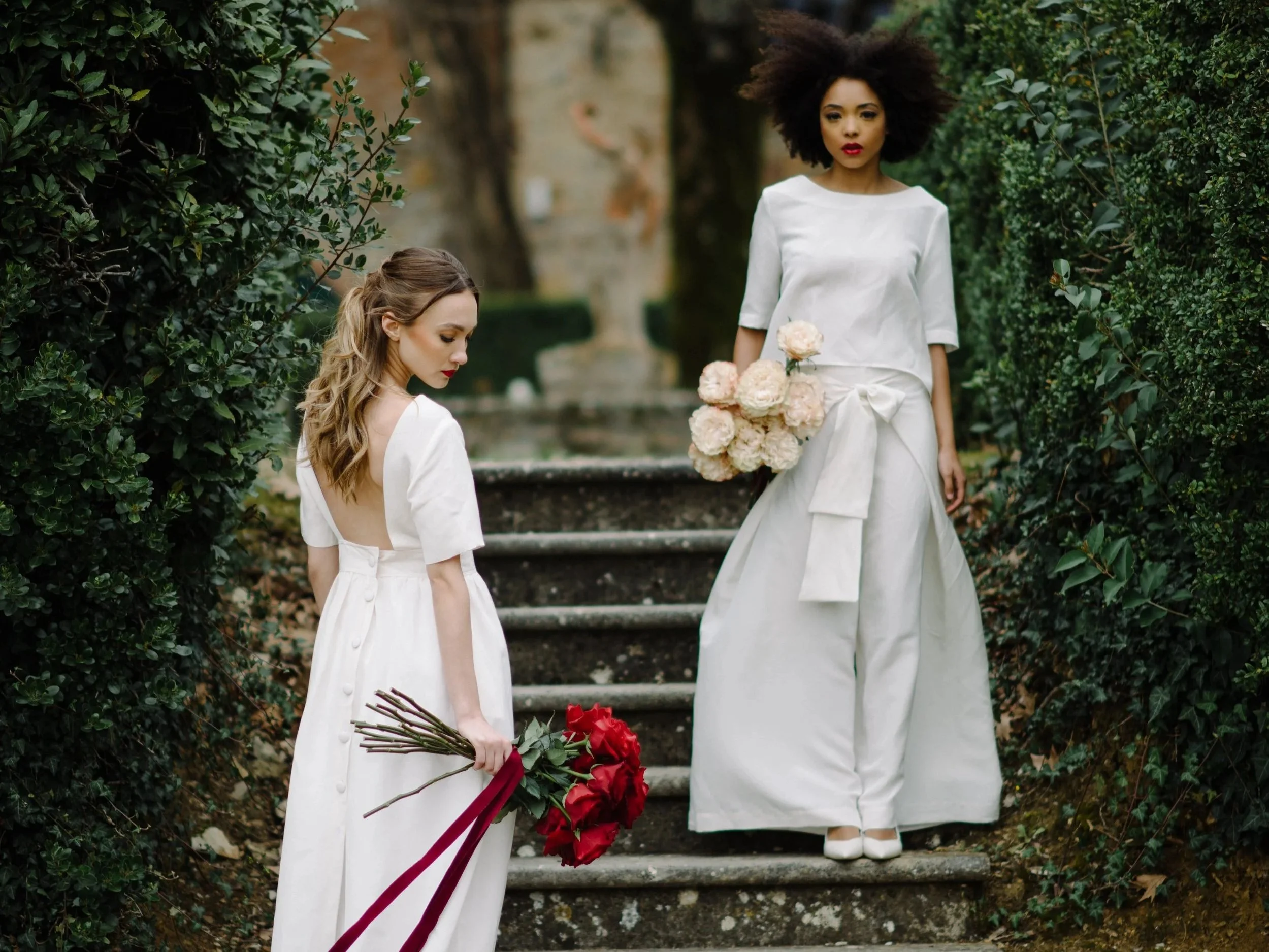 Two brides holding a long stemmed rose bouquet, one red and one white, at villa Nemora