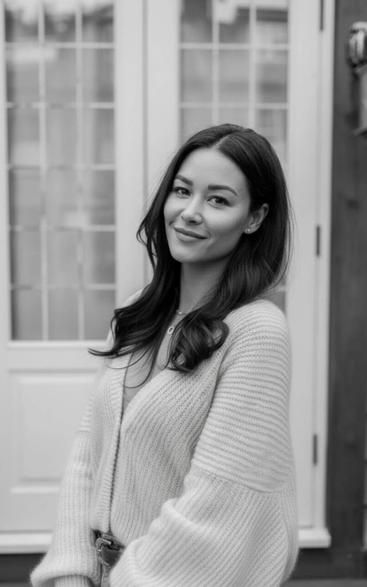 Black and white portrait of a woman with long, dark hair, smiling softly, wearing a cozy sweater, standing in front of a door with glass panes.