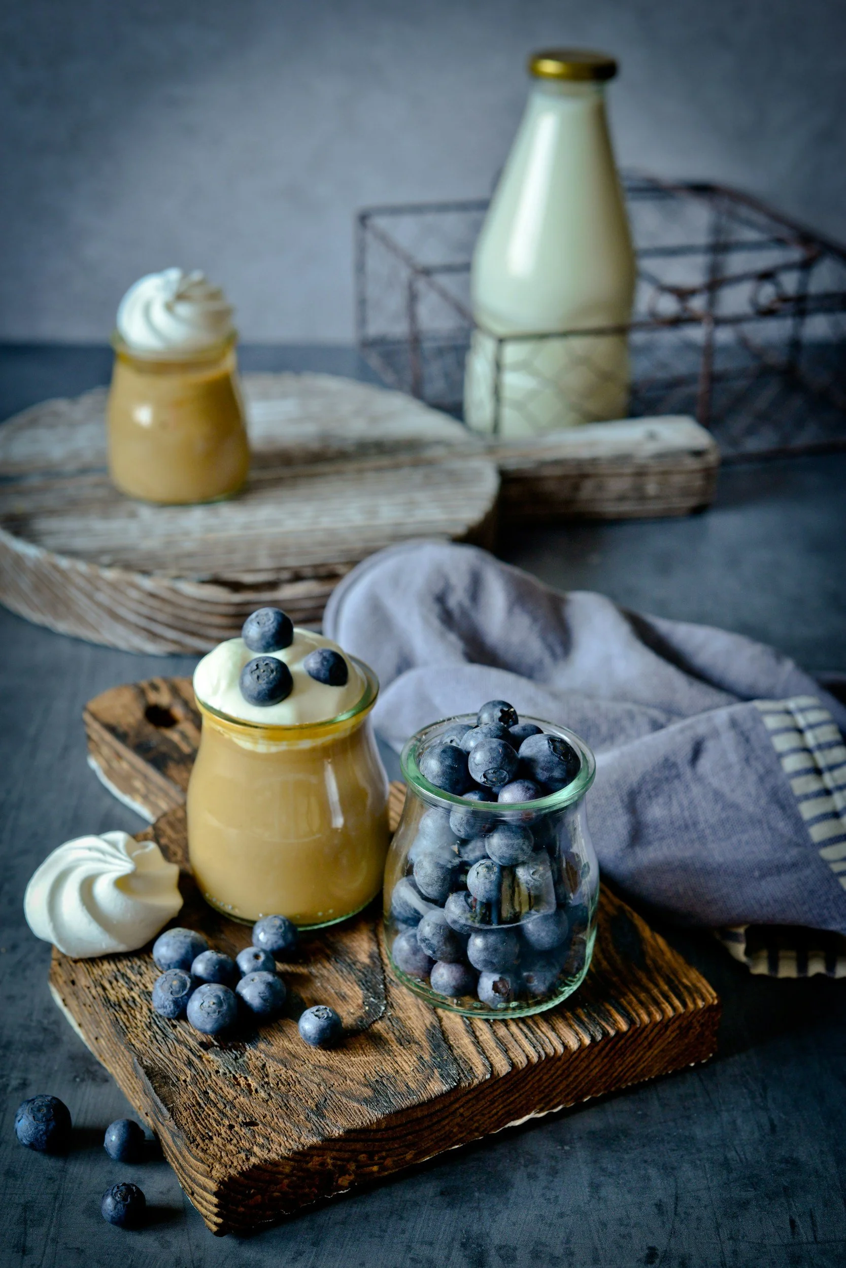 Small jars of blueberry-flavored dessert with blueberries on top and around, placed on rustic wooden boards, with a bottle of milk and a jar of blueberry compote in the background.