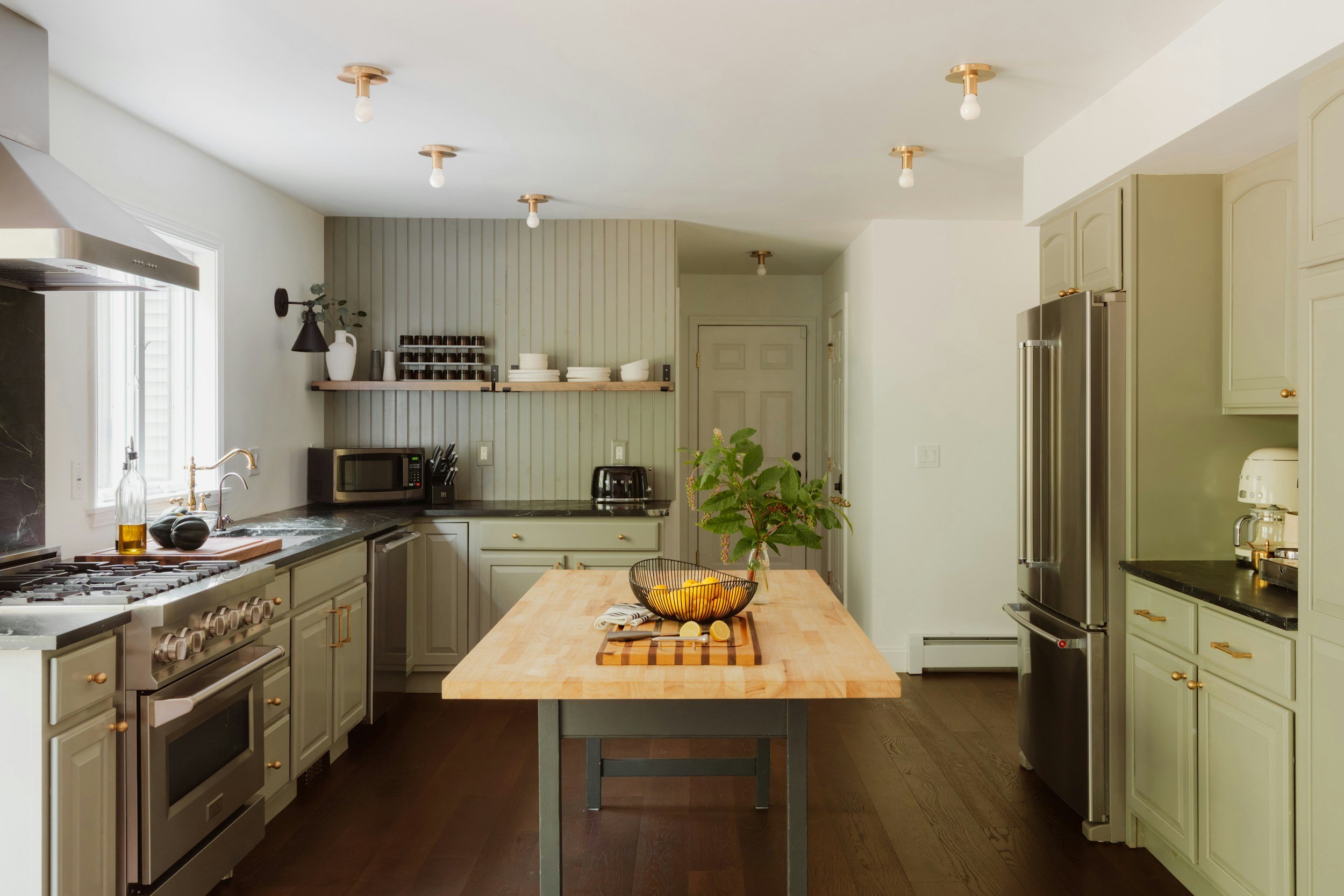 Modern kitchen with light green cabinets, stainless steel refrigerator, wooden island with greenery and bowl of lemons, dark hardwood floor, and various appliances and decor.