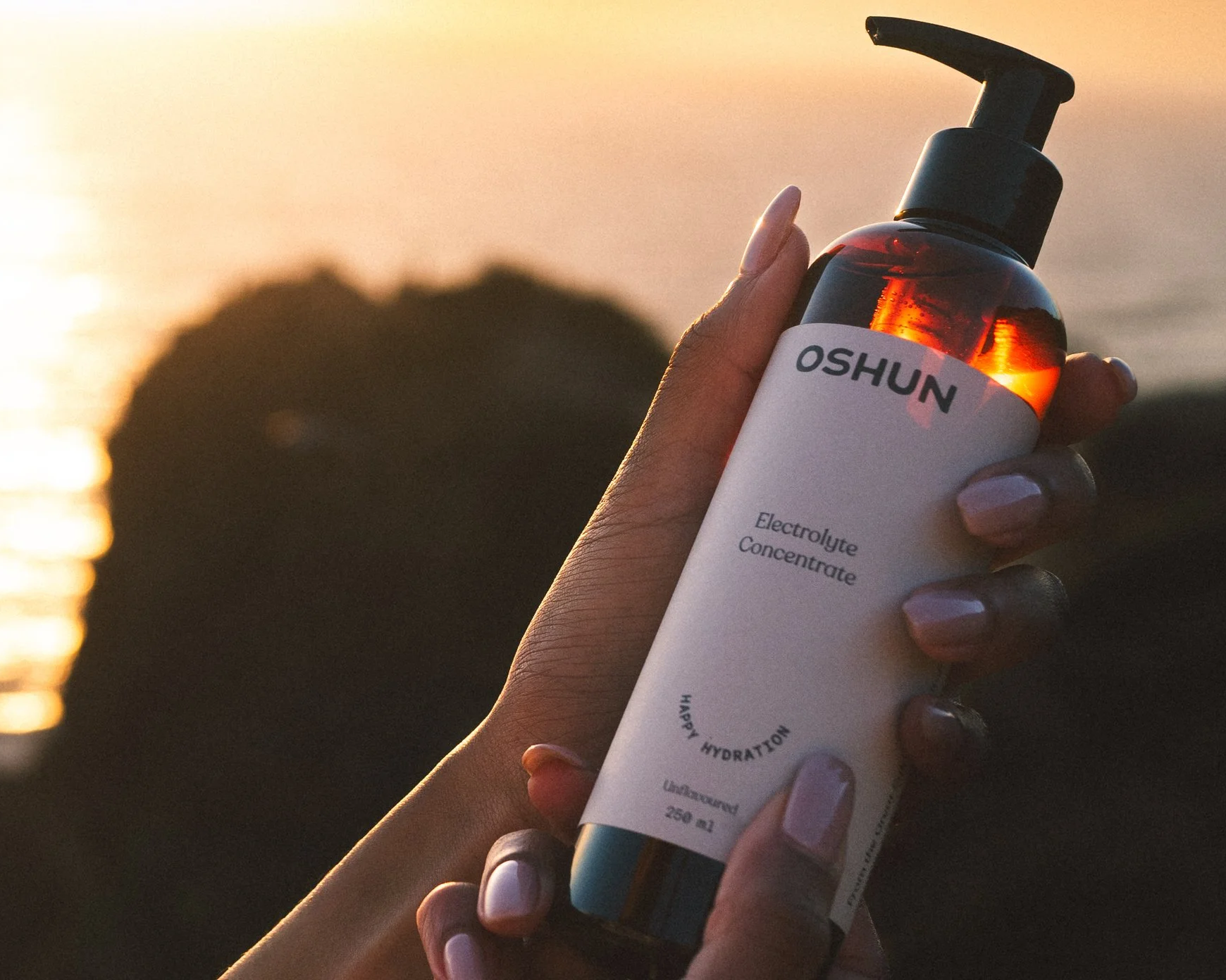 Hand holding a bottle of electrolyte concentrate labeled 'OSHUN' with sunset in the background.