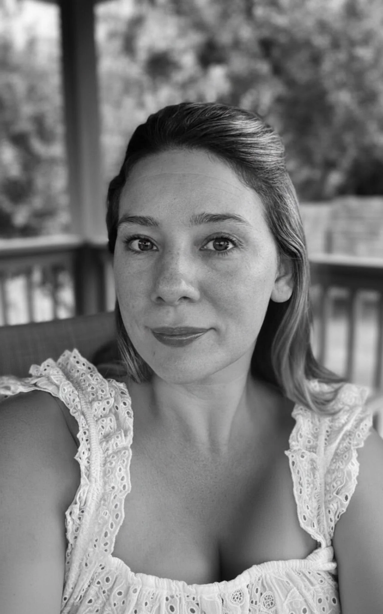 Black and white portrait of a woman with shoulder-length hair and a slight smile, wearing a lace-trimmed top, outside with blurred trees in the background.