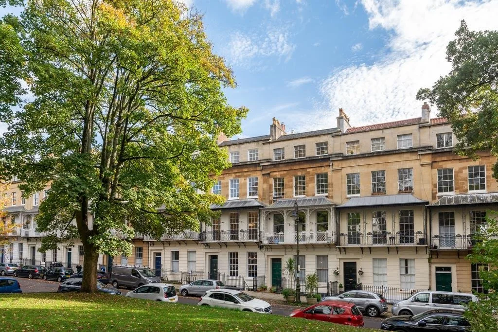 A row of multi-story townhouses with balconies and large windows, set against a backdrop of a partly cloudy sky and surrounded by green trees and parked cars.