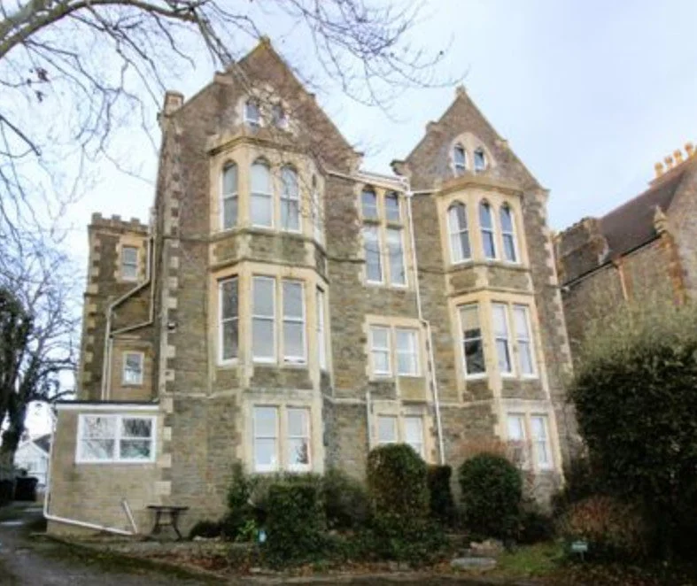 A large, old brick and stone multi-story residential building with several windows, surrounded by trees and bushes.