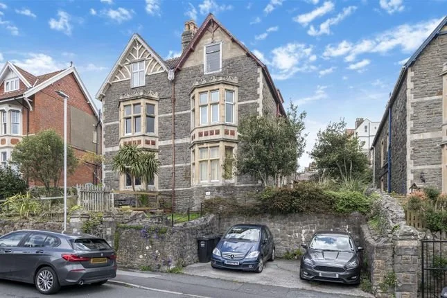 A large, historic stone house with decorative trim, tall windows, and multiple gables, situated on a hillside with trees, plants, and a small garden in front. Three cars are parked along the street in front of the house.