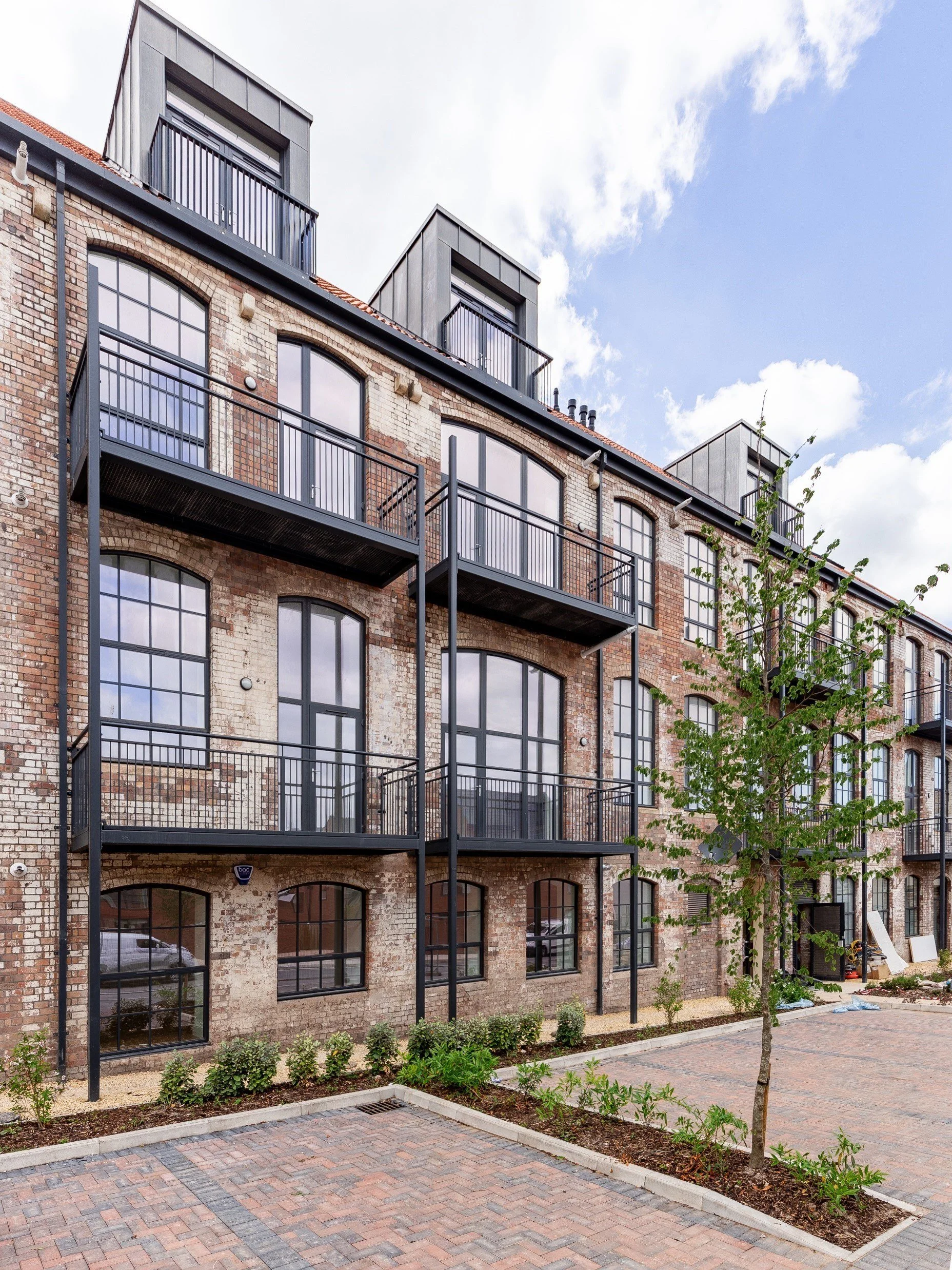 Modern brick apartment building with large windows and black metal balconies, a tree, and paved walkway in foreground under a partly cloudy sky.