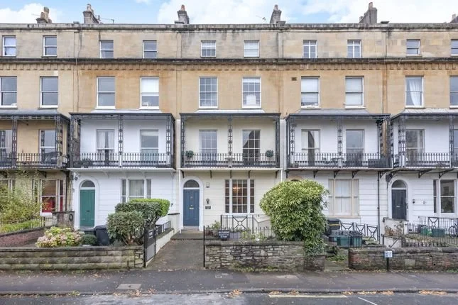A row of historic townhouses with cast iron balconies, some with plants or shrubs on the front steps, and a sidewalk with fallen leaves.