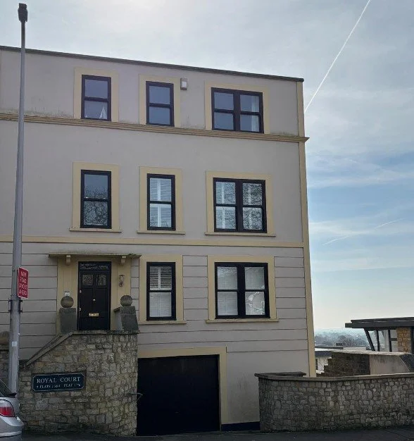 A multi-story beige building with black framed windows, a black front door with the number 3, and stairs with a stone wall leading to the entrance. There is a street sign that says 'Royal Court' and part of a parked car is visible on the left.
