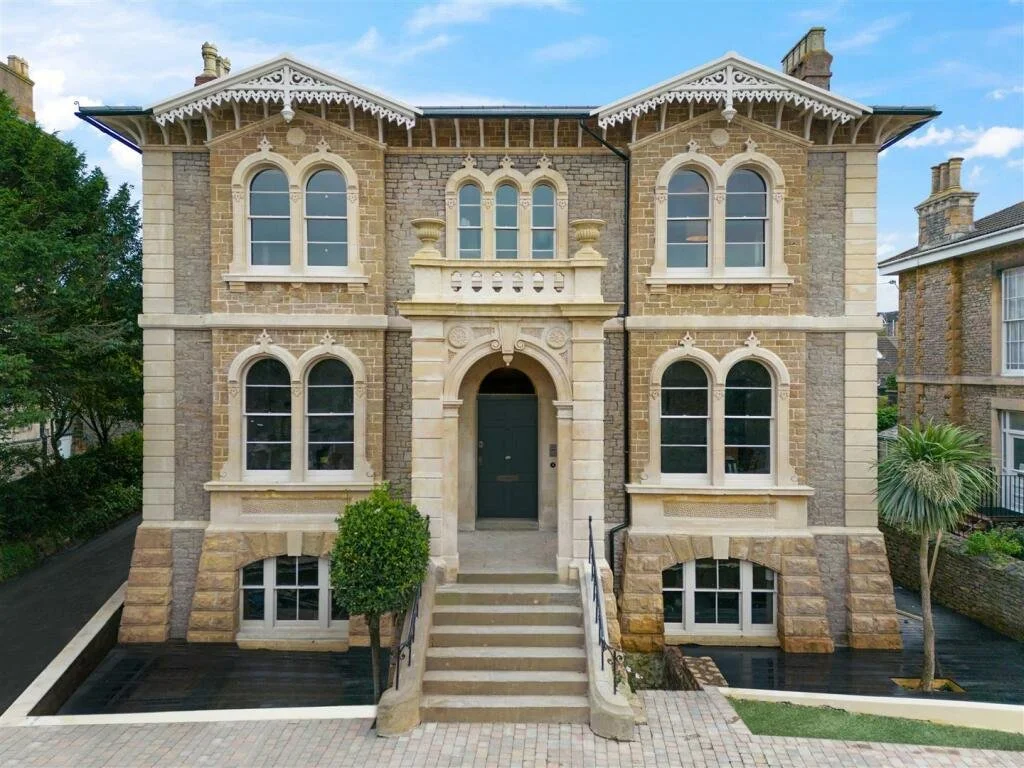 A large, historic brick house with ornate white trim, arched windows, and a central doorway accessed by stairs with black railings. There are trees and neighboring houses nearby.