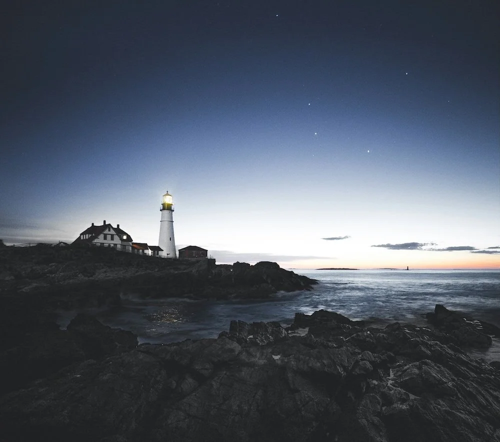 a lighthouse shines its beam across the water at dusk as stars appear in the sky