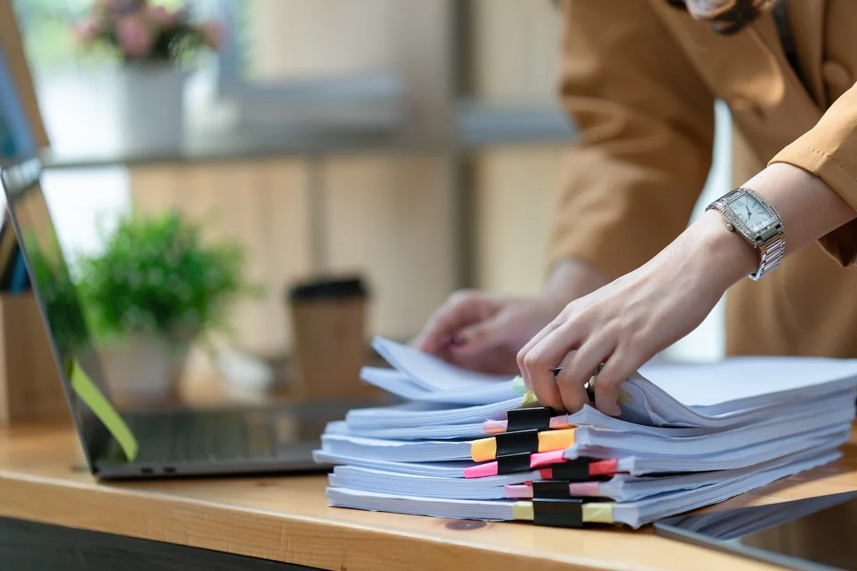 woman's hand flipping through a stack of bundled documents with a laptop in the background