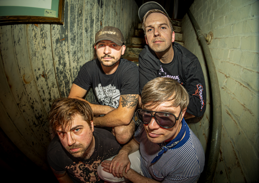 Four men posing in a small, worn staircase or corner of a building with wooden and brick walls.