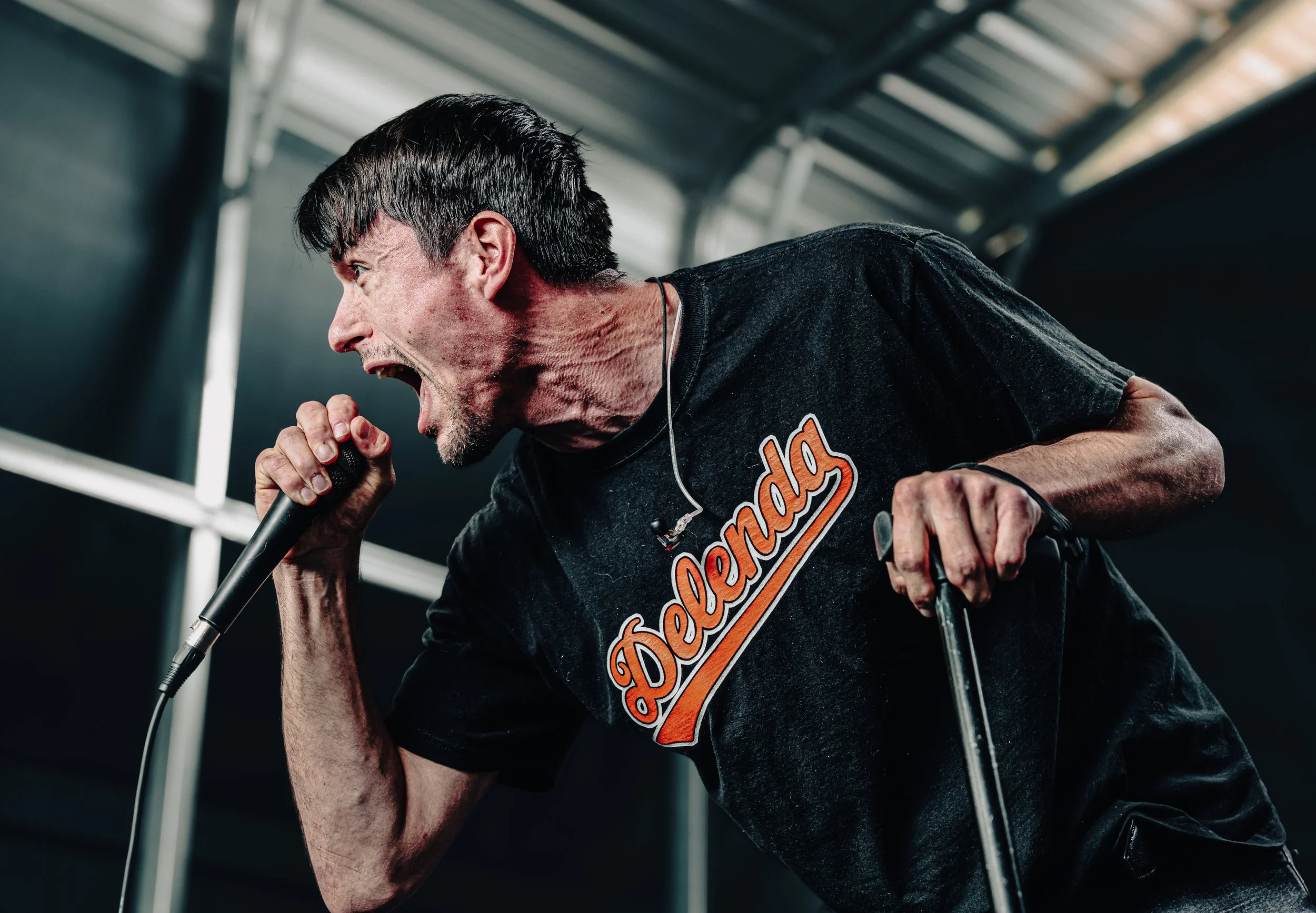 A male singer passionately performing on stage, holding a microphone with his left hand, wearing a black T-shirt with the word 'Dedlands' written on it.