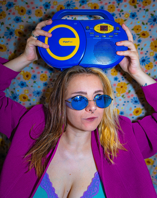 Woman with long hair, wearing blue sunglasses and a purple jacket, holding a blue boombox on her head, standing in front of a floral patterned background.