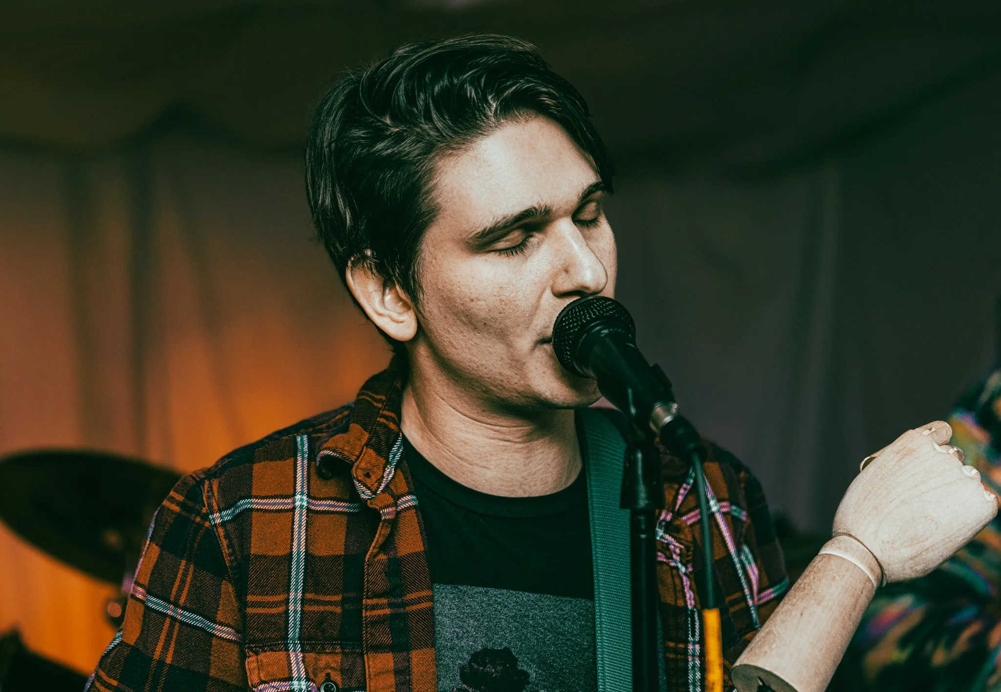 A young man with dark hair singing into a microphone, wearing a red and black plaid shirt over a graphic T-shirt, with eyes closed and a wooden arm and hand prop on his left.