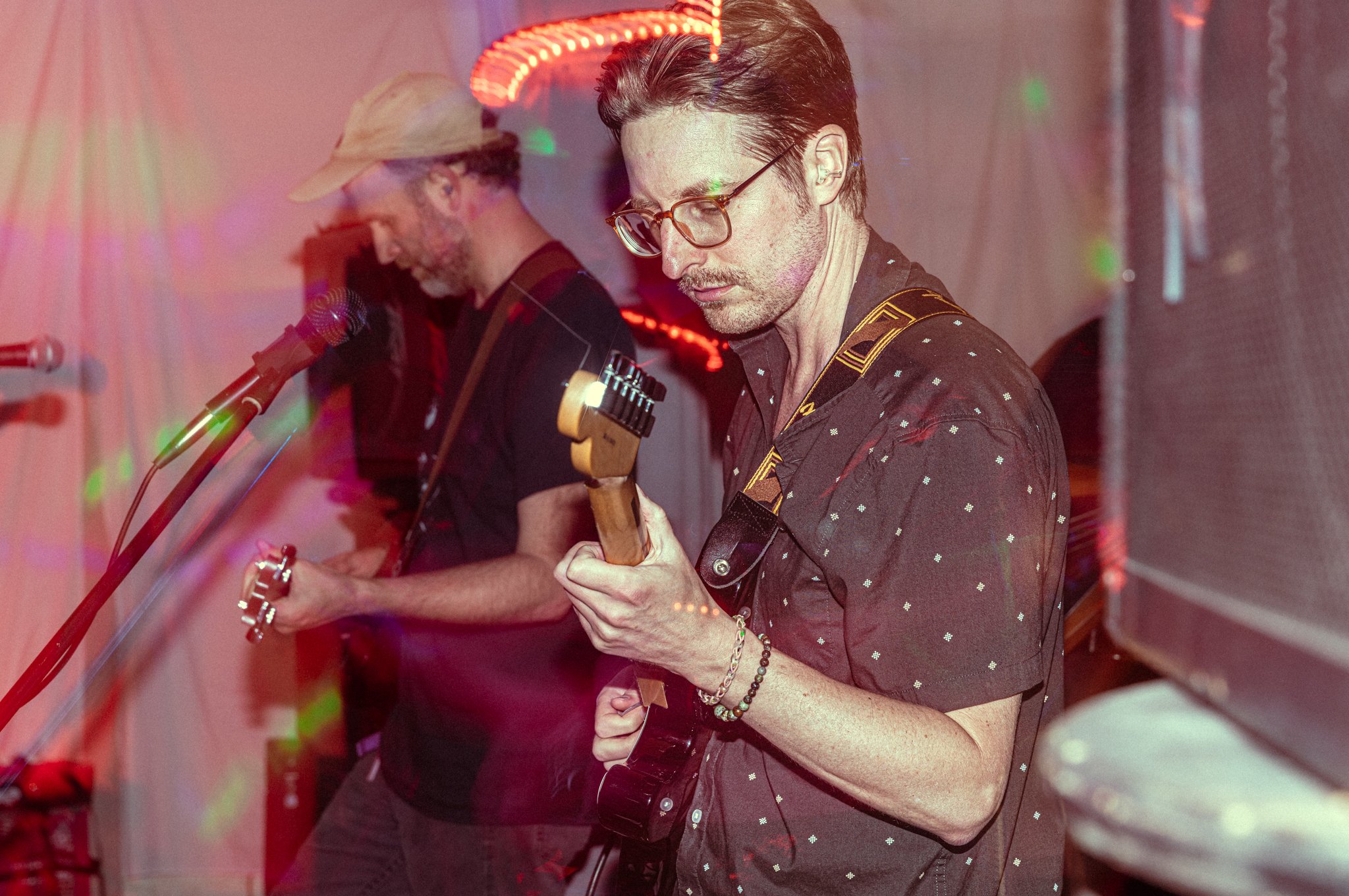Two musicians performing, one playing guitar in the foreground and the other singing or playing an instrument in the background, under colorful stage lighting.