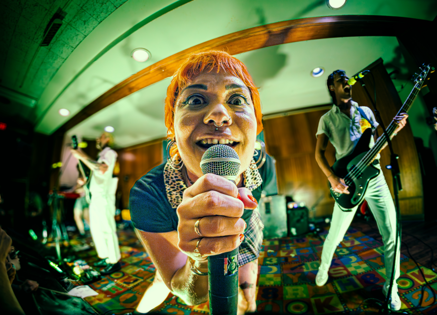 Person with short orange hair holding a microphone, performing on stage with band members playing guitar and keyboard in background, colorful patterned floor.