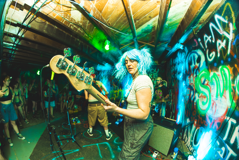 A woman with blue, curly hair playing bass guitar on stage at a lively underground music venue with graffiti-covered walls and an audience of young people watching.