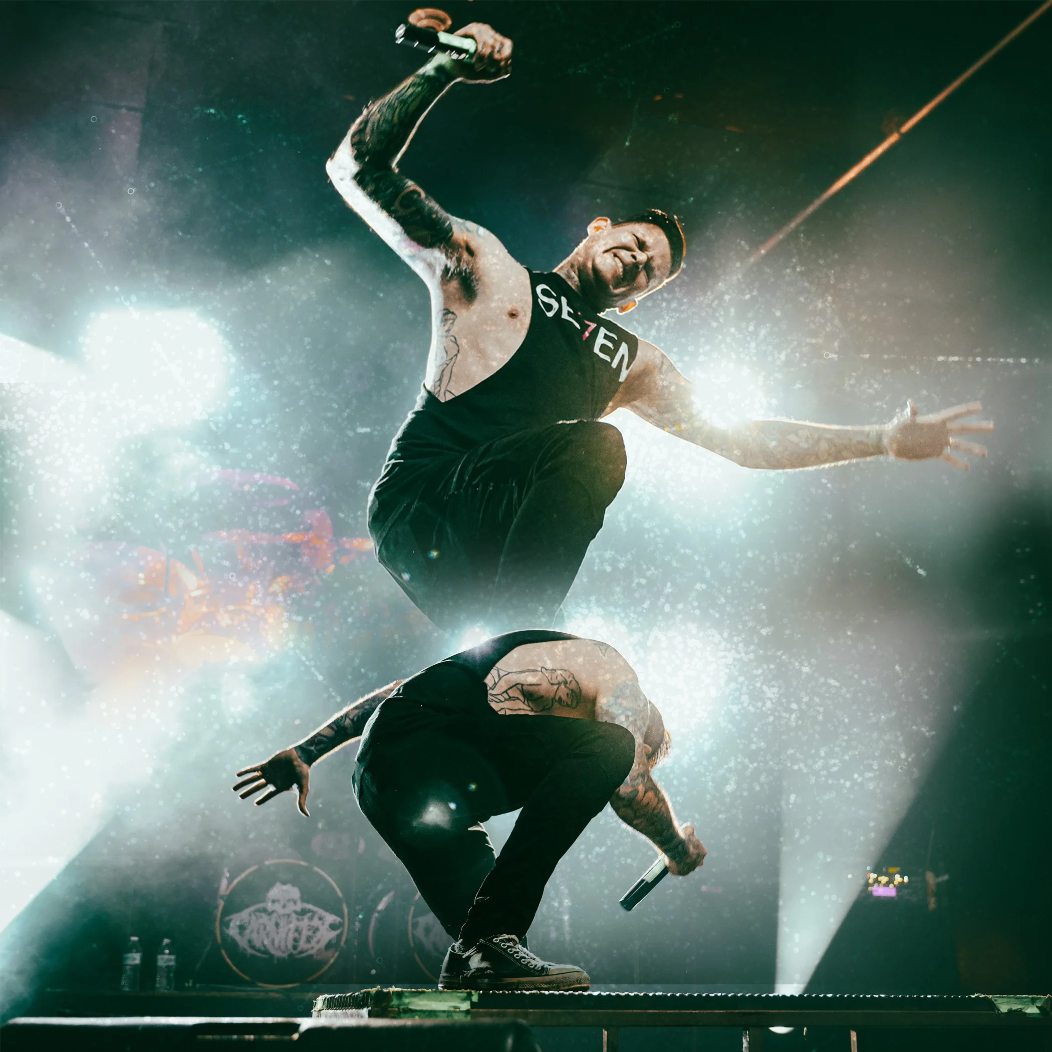 Two tattooed male performers on stage, one squatting and holding a microphone, the other jumping with a microphone in hand, surrounded by bright stage lights and fog, during a concert.