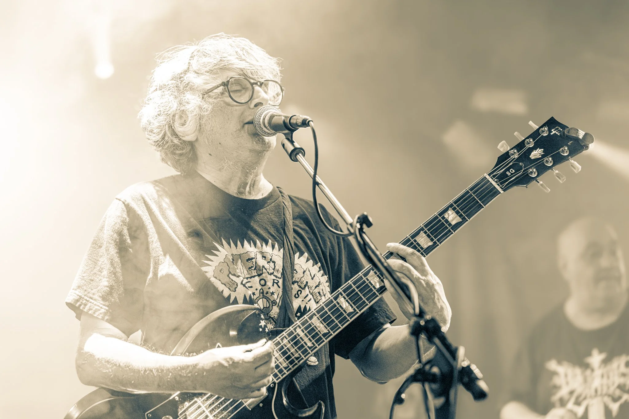An elderly man with curly gray hair and glasses playing an acoustic guitar and singing into a microphone during a live performance, with another person in the background.