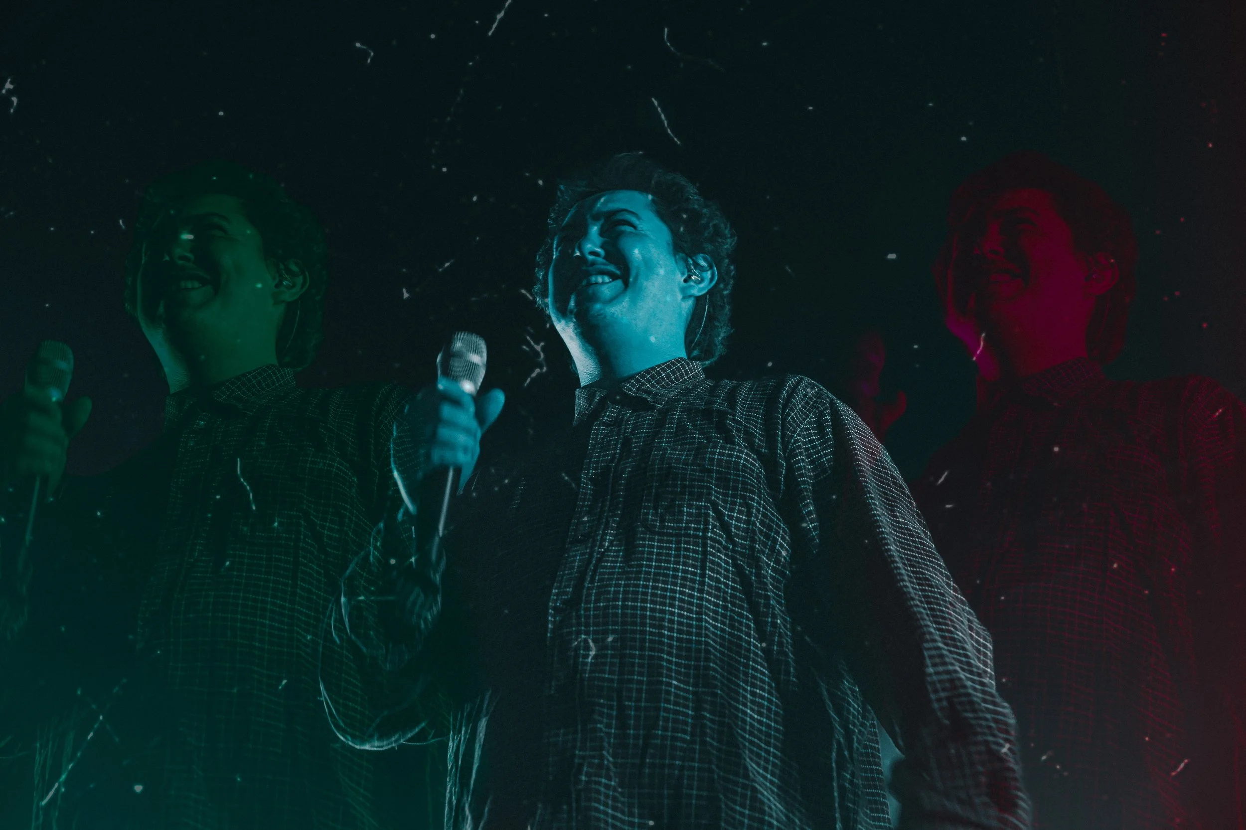 Three smiling women on stage holding microphones, illuminated with colored lights, under dark background with specks of light or confetti.