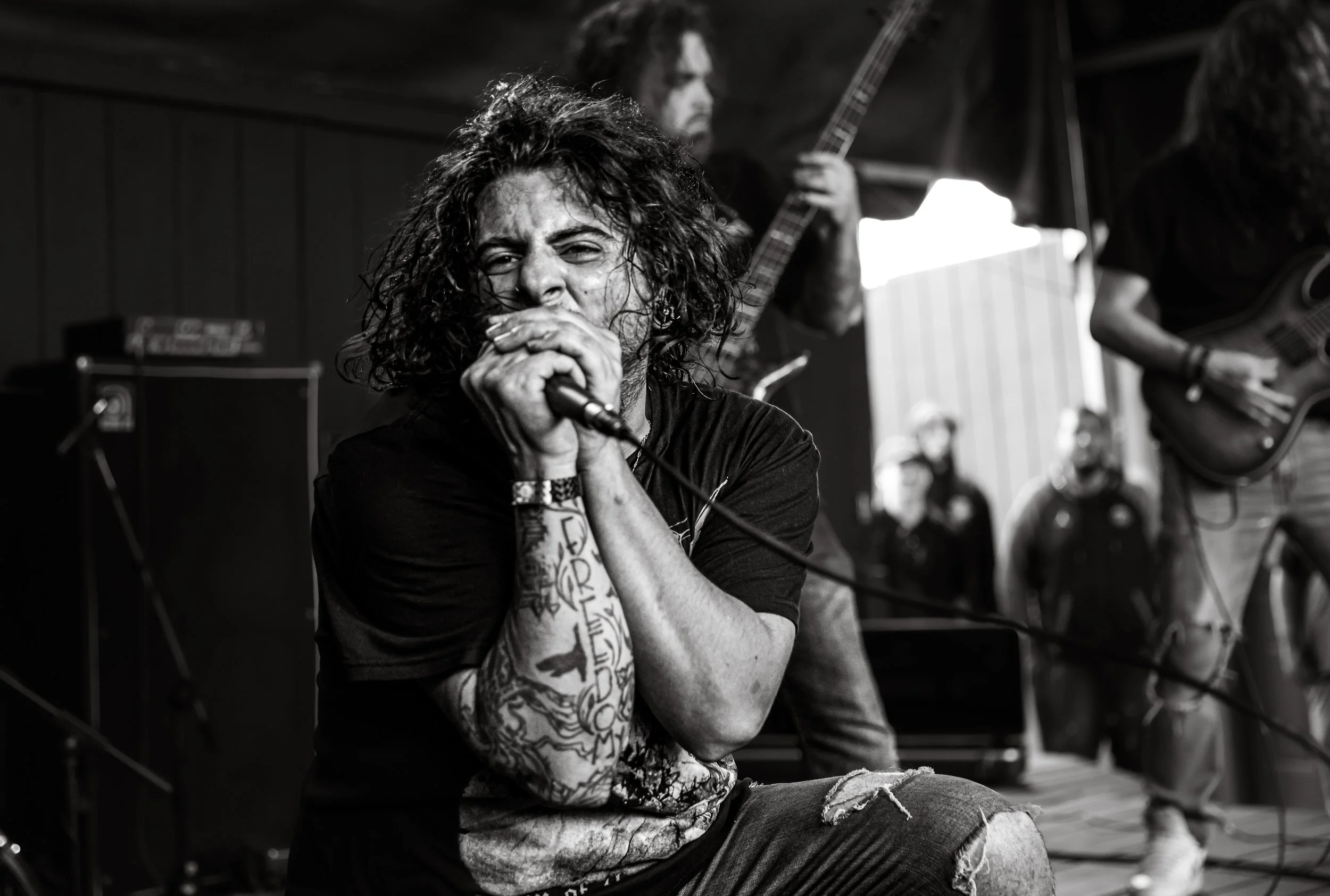 Black and white photo of a punk rock singer with curly hair, tattoos on their arm, and torn jeans, holding a microphone close to their mouth during a performance. Two musicians playing guitars are visible in the background.