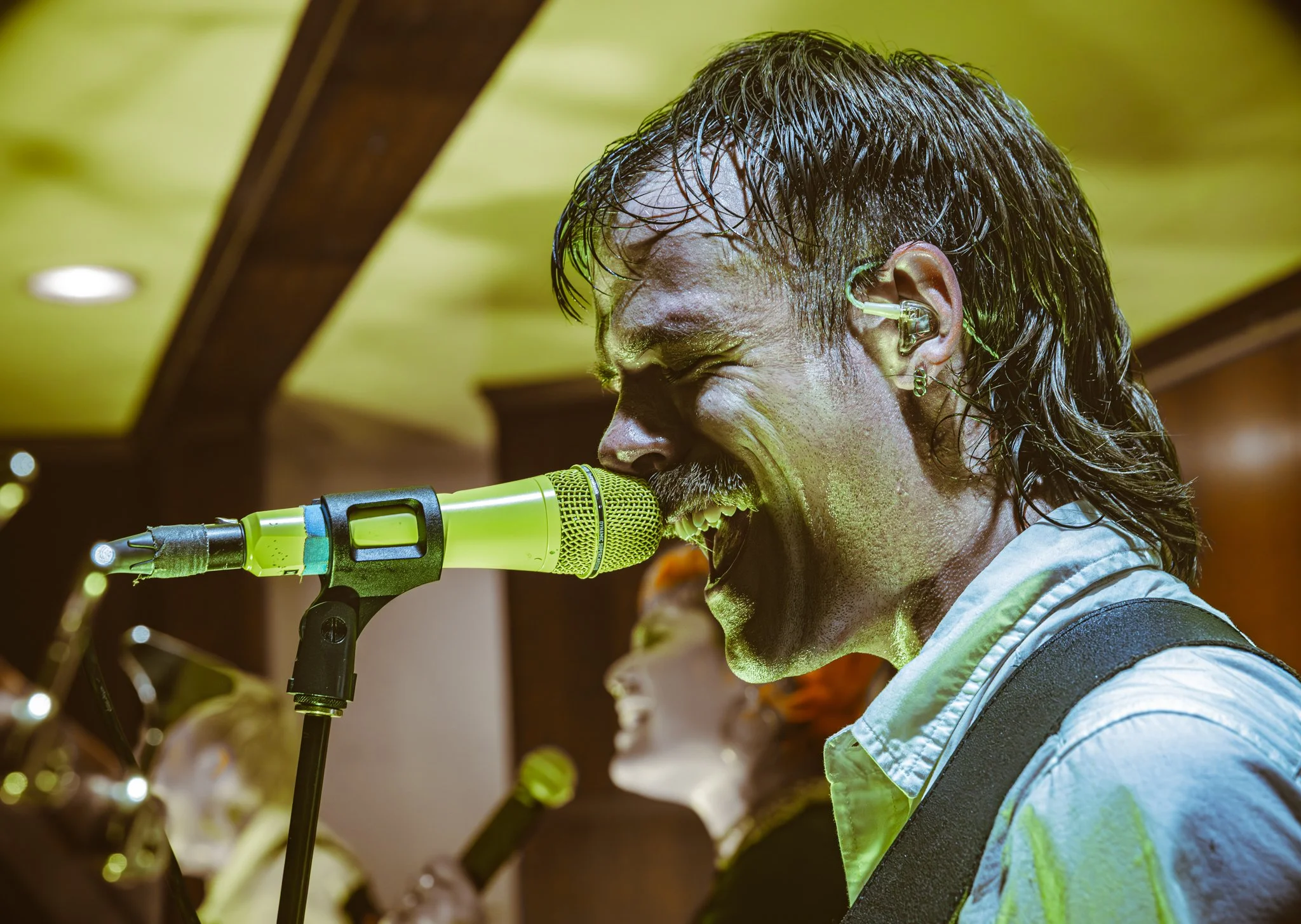 A male musician singing passionately into a green microphone on stage, with wet long dark hair and wearing an earpiece and a light-colored shirt.