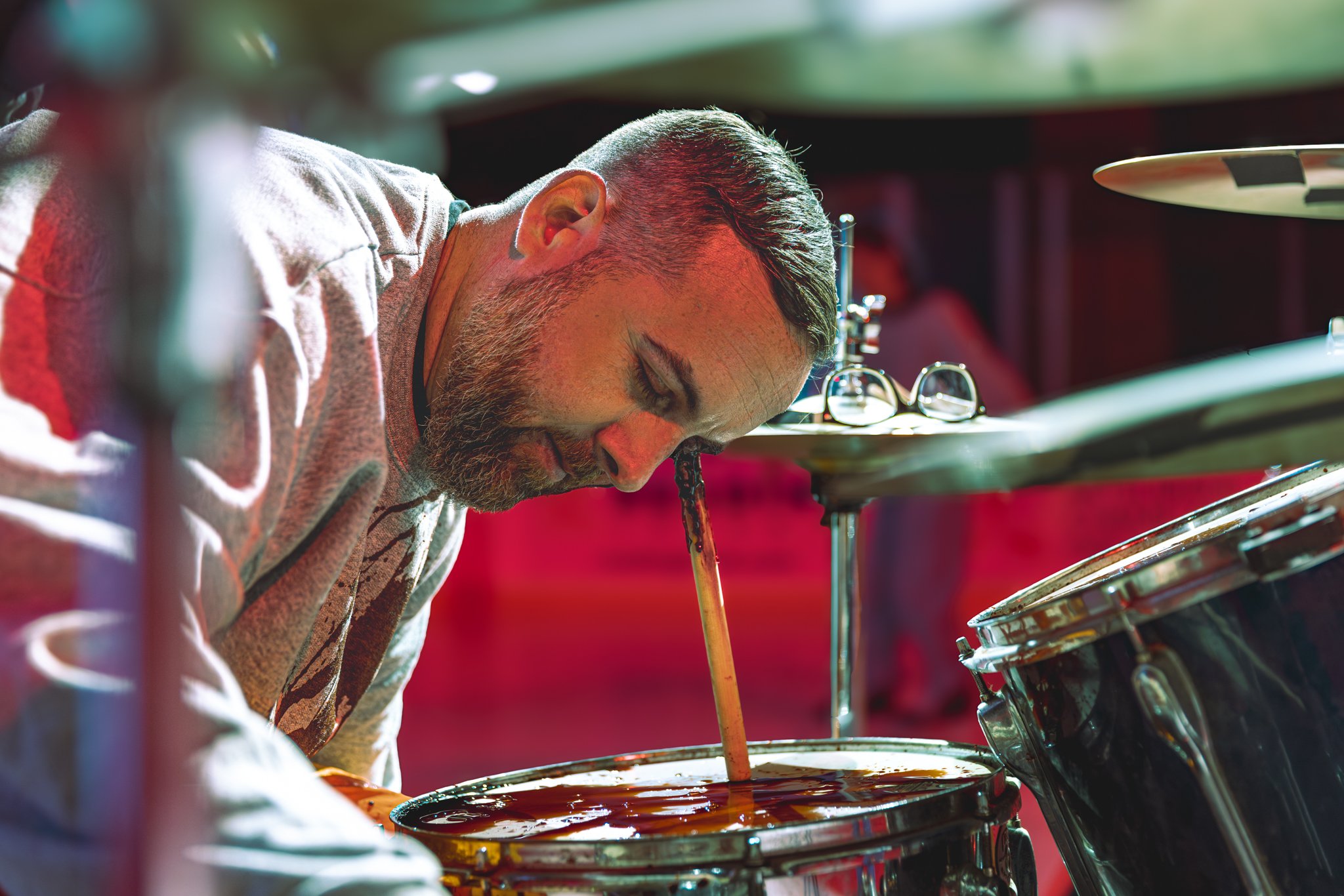 Musician with short hair and beard playing drums with drumsticks, glasses resting on a drum, in a dimly lit setting with red and green lighting.