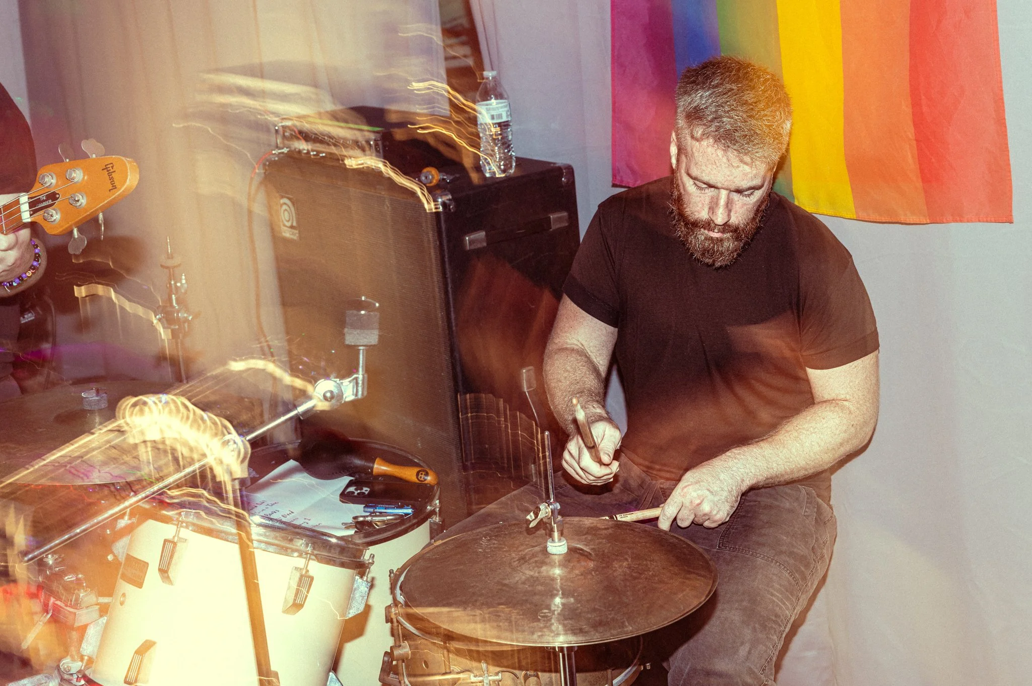 A man playing drums at a music event with a rainbow pride flag on the wall behind him.