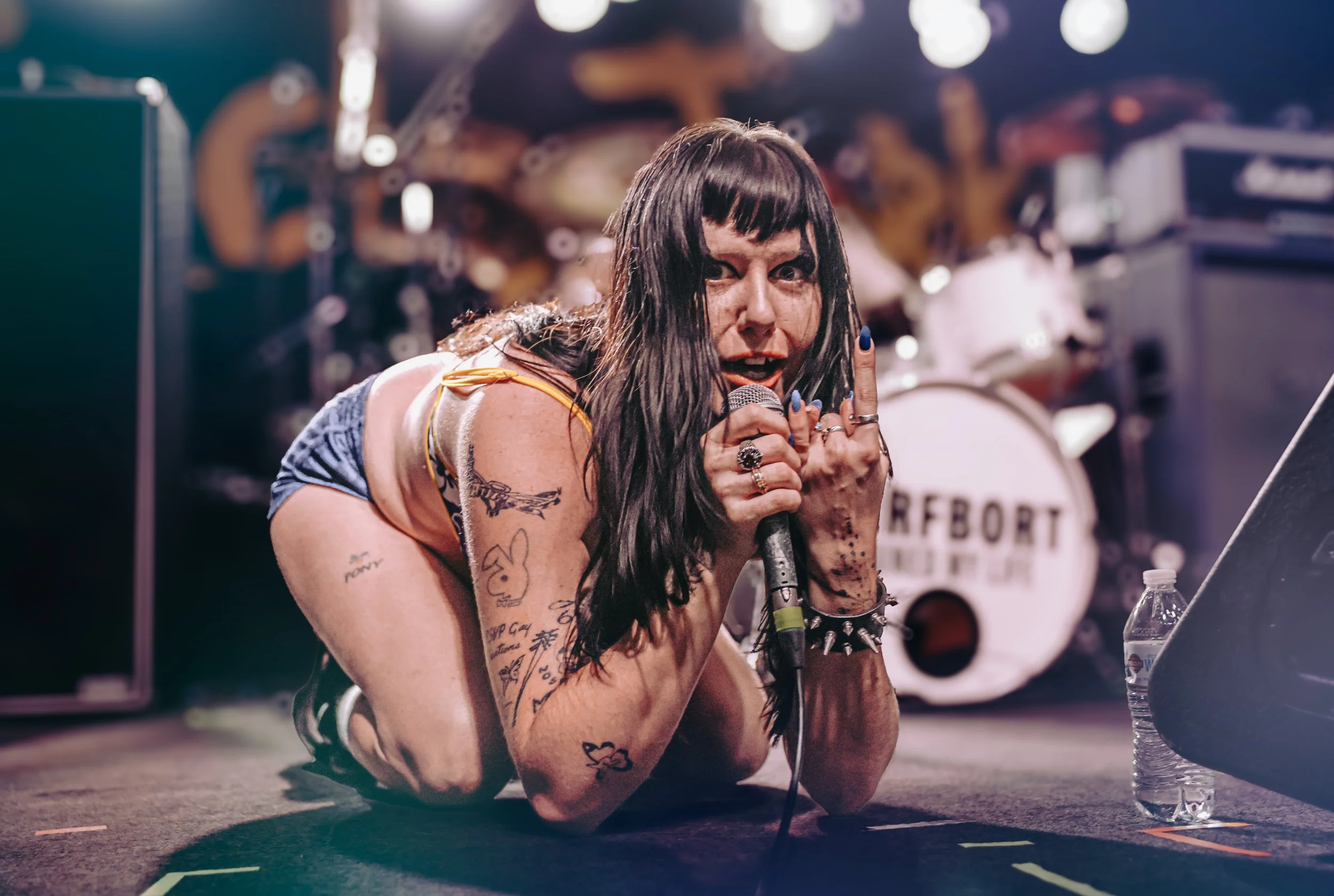 A woman with long dark hair, tattoos on her arms, and dark eye makeup is lying on the stage floor, holding a microphone and looking intensely at the camera during a live music performance.