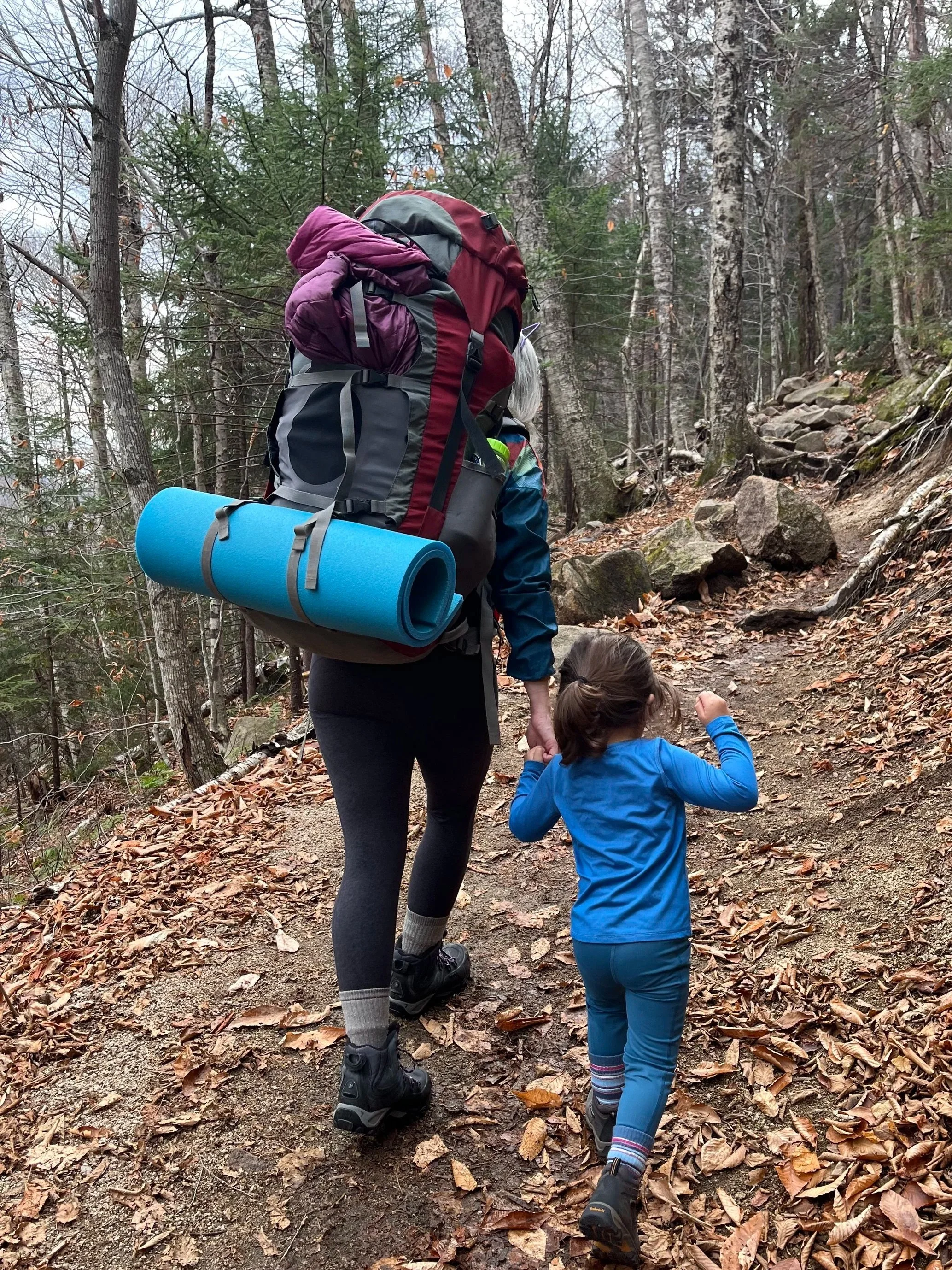 A person carrying a child on a hike in a forest with trees and a rocky trail, both wearing hiking boots, surrounded by fallen leaves.