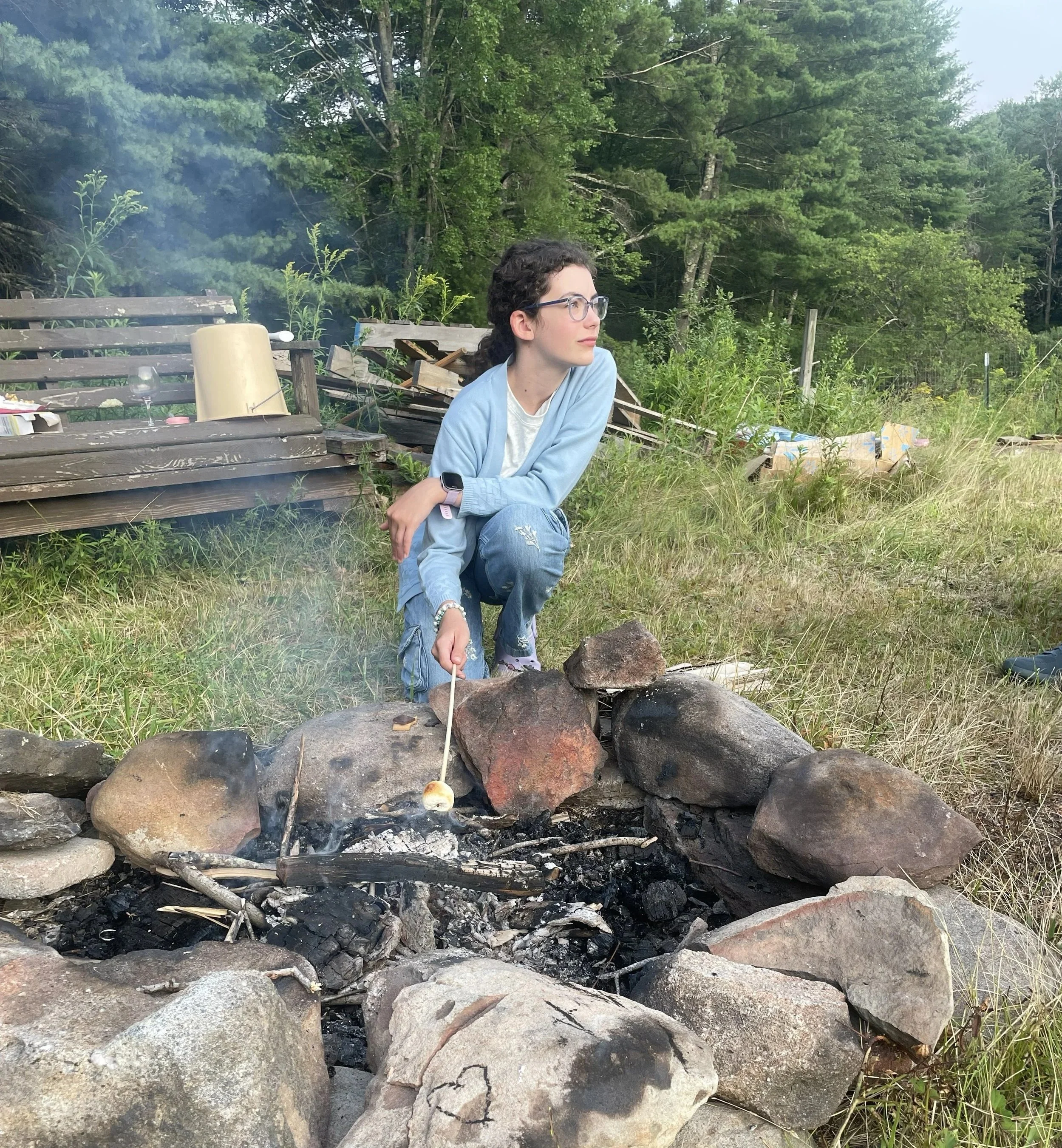 A woman with curly dark hair, glasses, wearing a light blue jacket, is sitting by a small campfire made of rocks, in a grassy outdoor area with trees in the background. Smoke rises from the fire, and there are items like a wine glass and some paper on a wooden bench nearby.