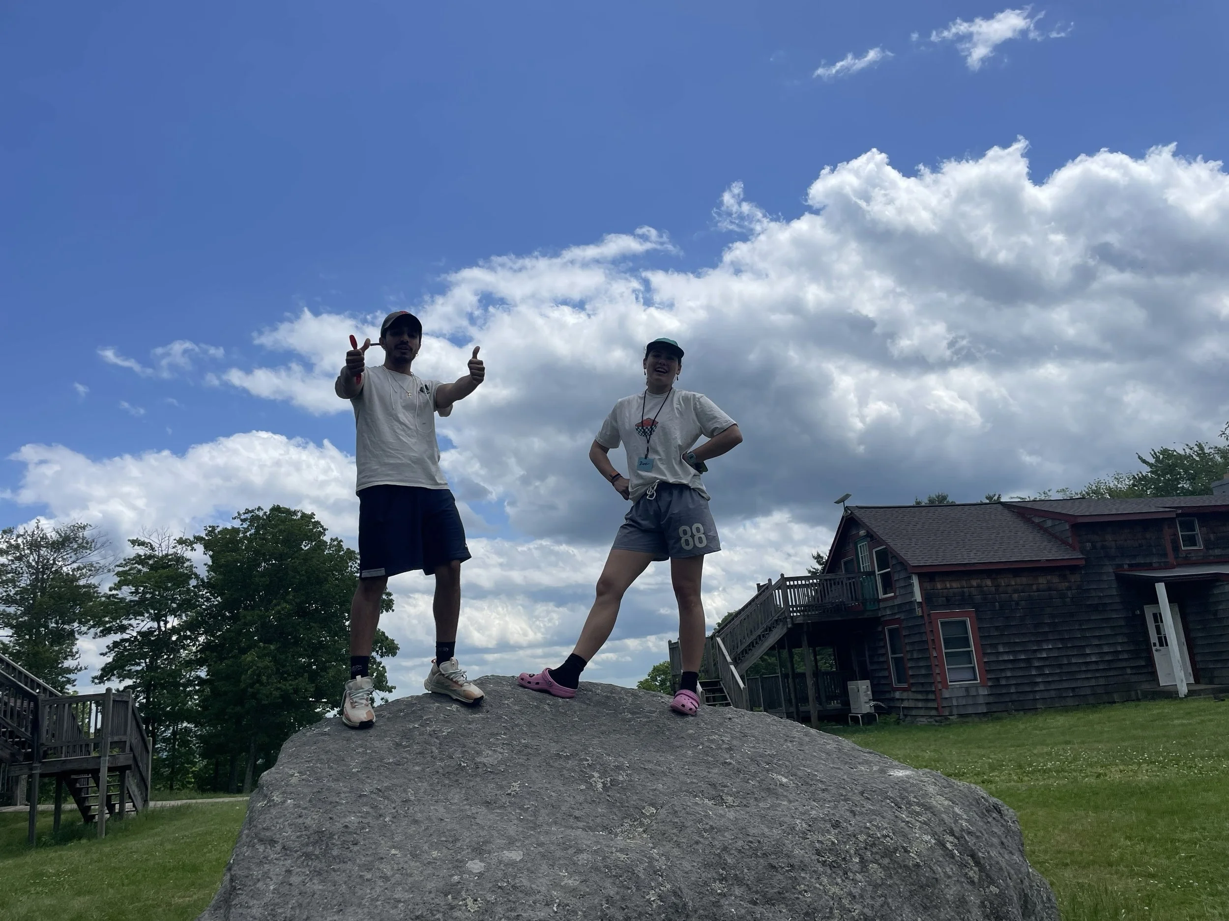 Two young people standing on top of a large rock outdoors on a grassy area, with a house and trees in the background. The person on the left is giving a thumbs-up with both hands, and the person on the right is standing with one hand on their waist, both smiling under a partly cloudy sky.
