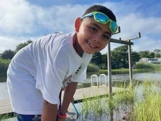 A boy smiling at the camera outdoors near a pond, wearing goggles and a white t-shirt.