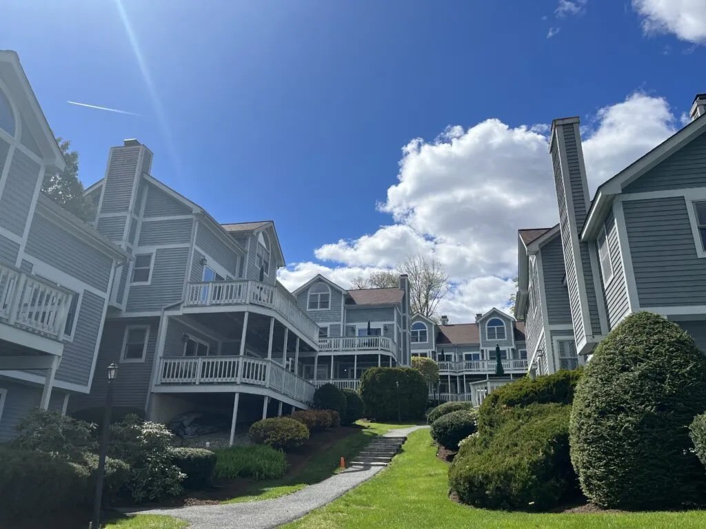  A view looking uphill at many of the rental building. The buildings are blue with white trim. Green grass, shrubs and a blue sky are also visible. 