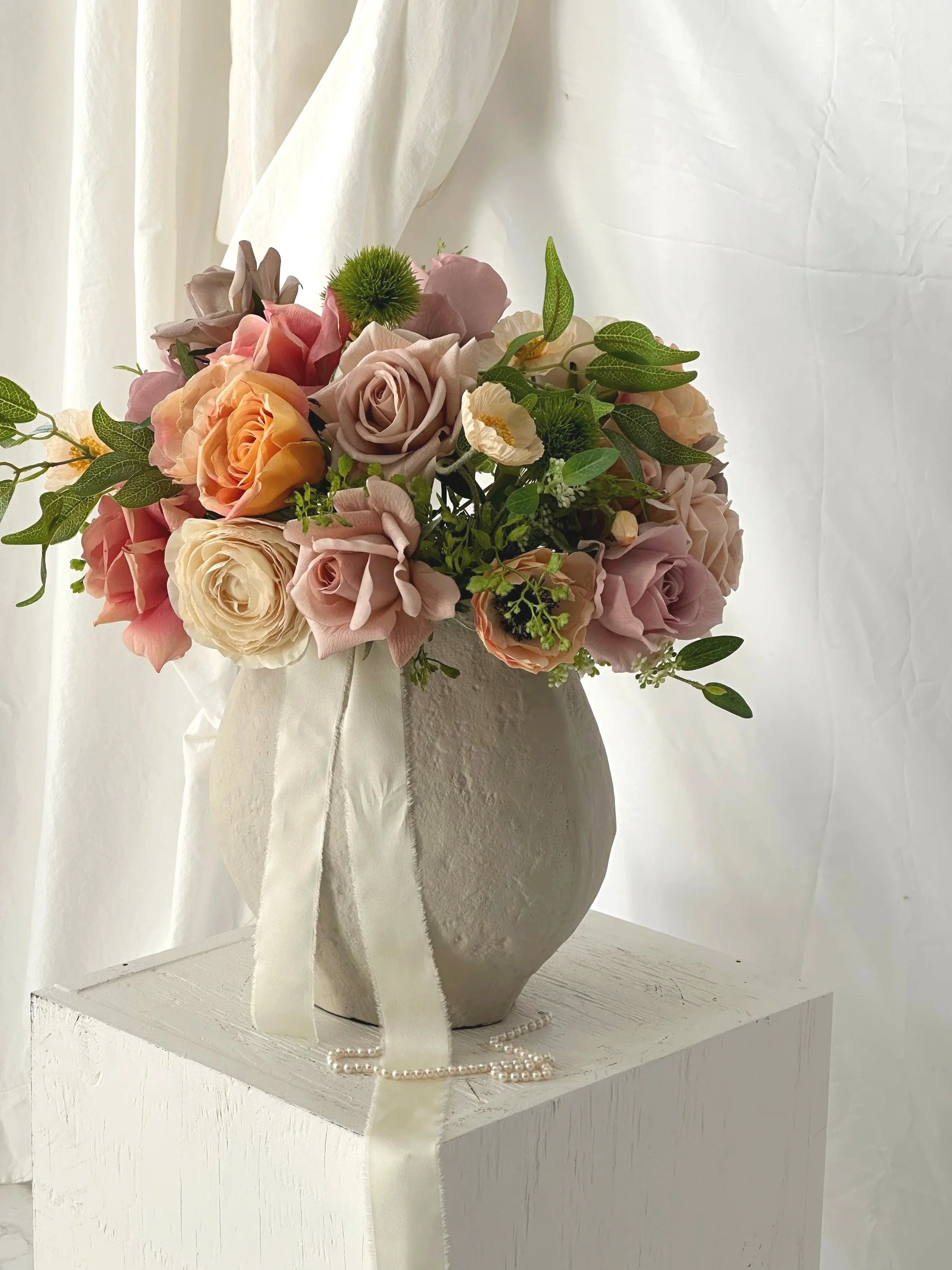 A large bridal bouquet with greenery and light pink, peach, white and lavender roses sits in a large organically made and styled clay urn on top of a pedestal