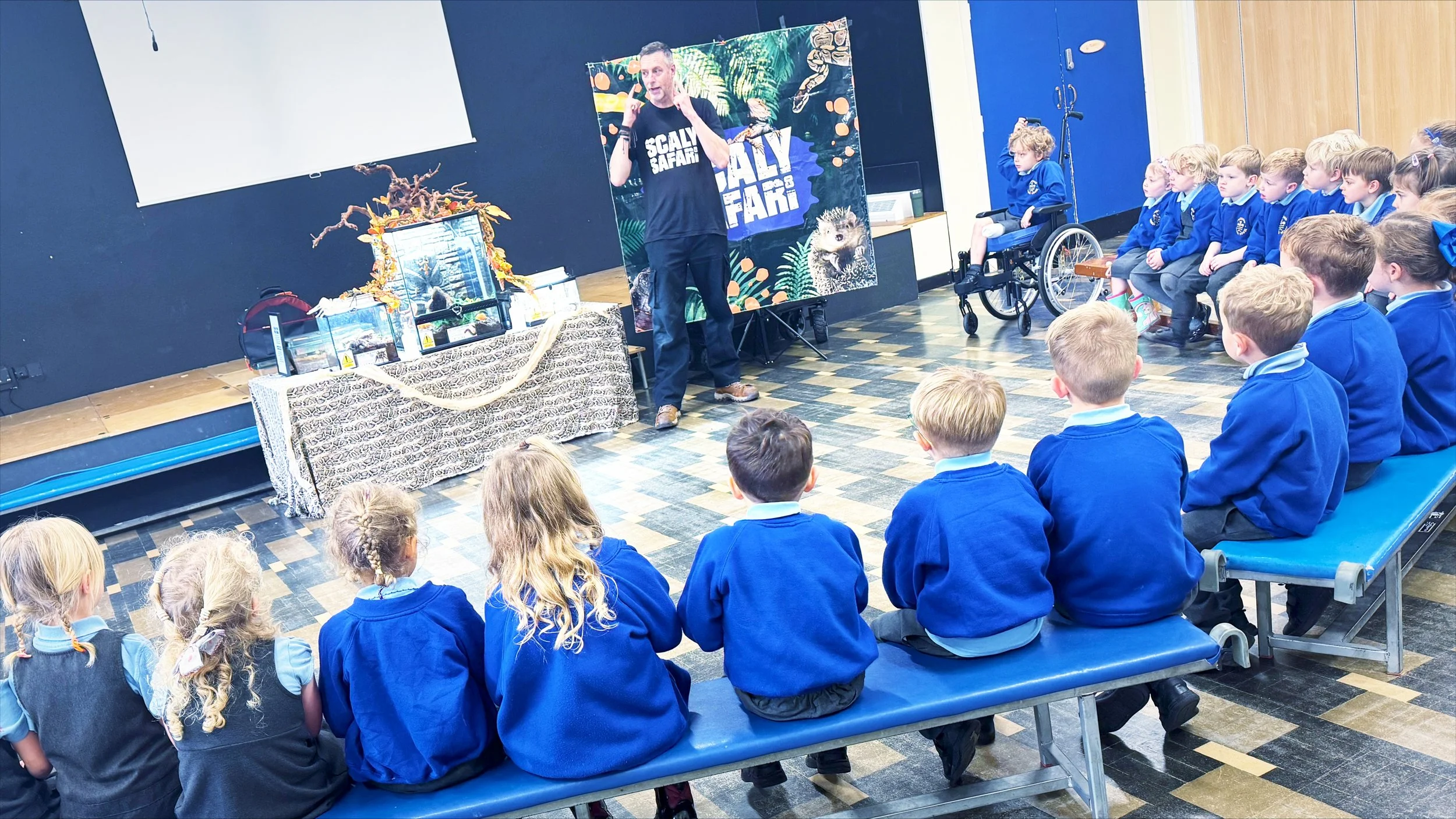 A Scaly Safari reptile workshop taking place in a school hall, with a presenter standing at the front while primary school children in blue uniforms sit on benches listening attentively.