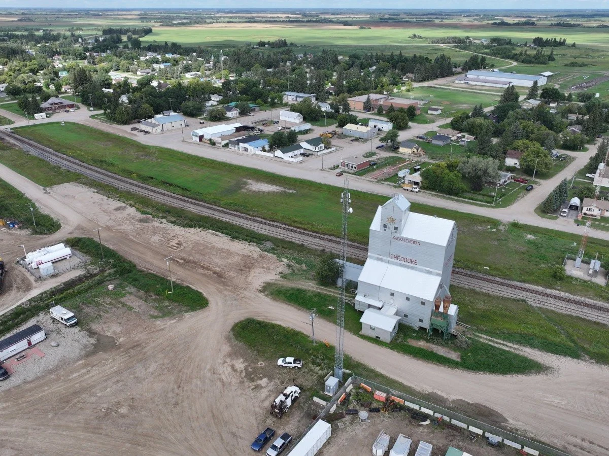 An aerial view of a small town with a grain elevator labeled "Saskatchewan" and "Theodore" in the foreground, railway tracks, and residential and industrial buildings surrounded by fields.