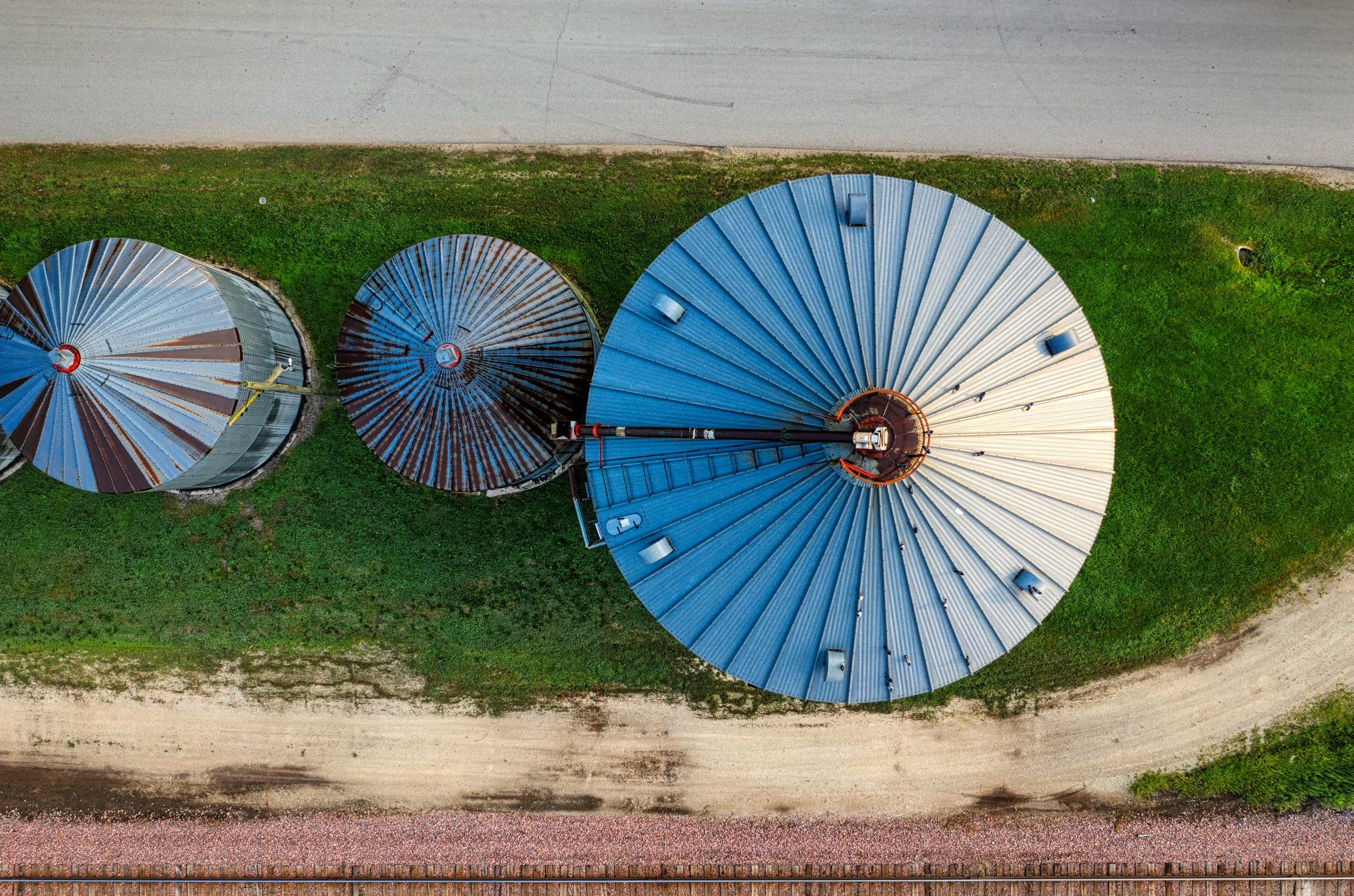 An aerial view showing four circular grain silos with corrugated metal roofs, situated on a green grassy area next to a dirt road and railway tracks.