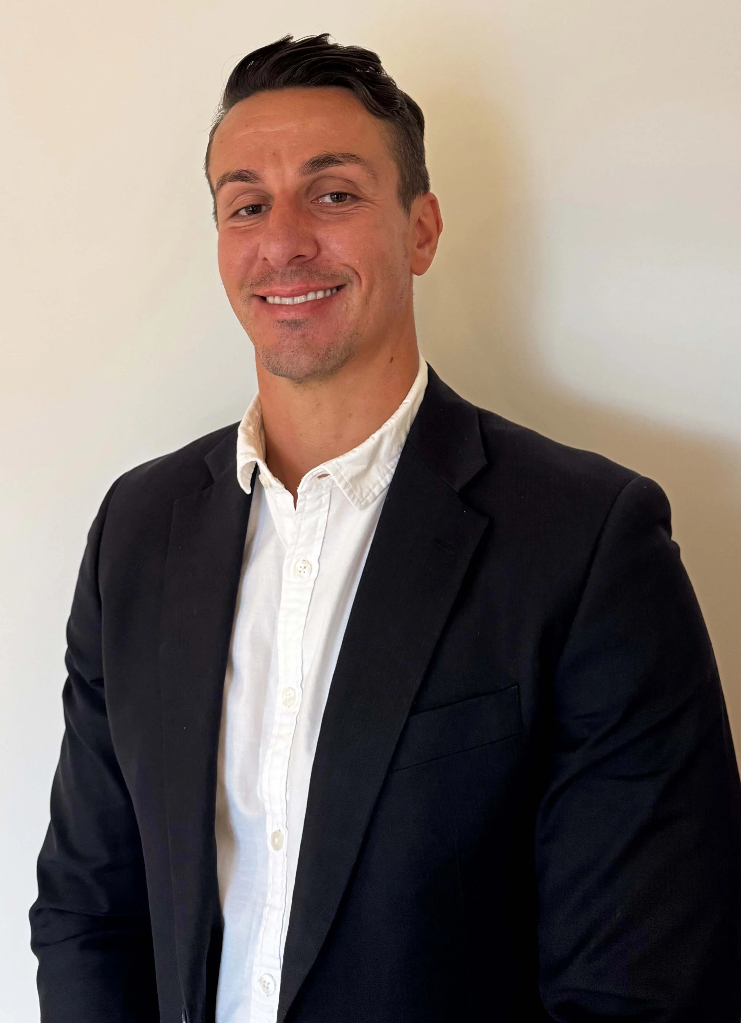 A young man in a black suit jacket and white dress shirt smiling against a plain, light-colored background.
