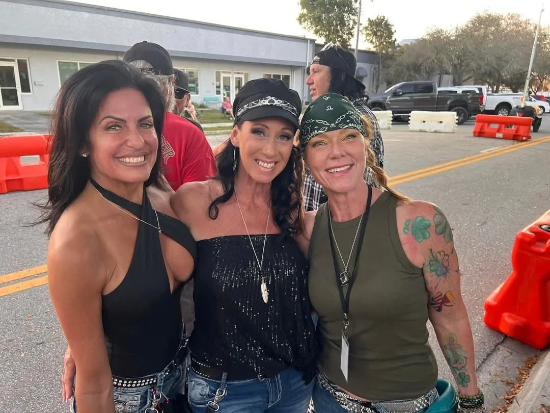 Three women smiling outdoors near a street with parked cars and buildings in the background. They are wearing casual and motorcycle-themed attire, some with bandanas and jewelry.