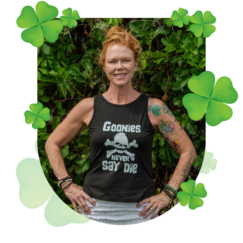 A woman with red hair and colorful tattoos wearing a black sleeveless shirt that says "Goonies Never Say Die". She is outside standing in front of green foliage, smiling with her hands on her hips, surrounded by shamrocks.