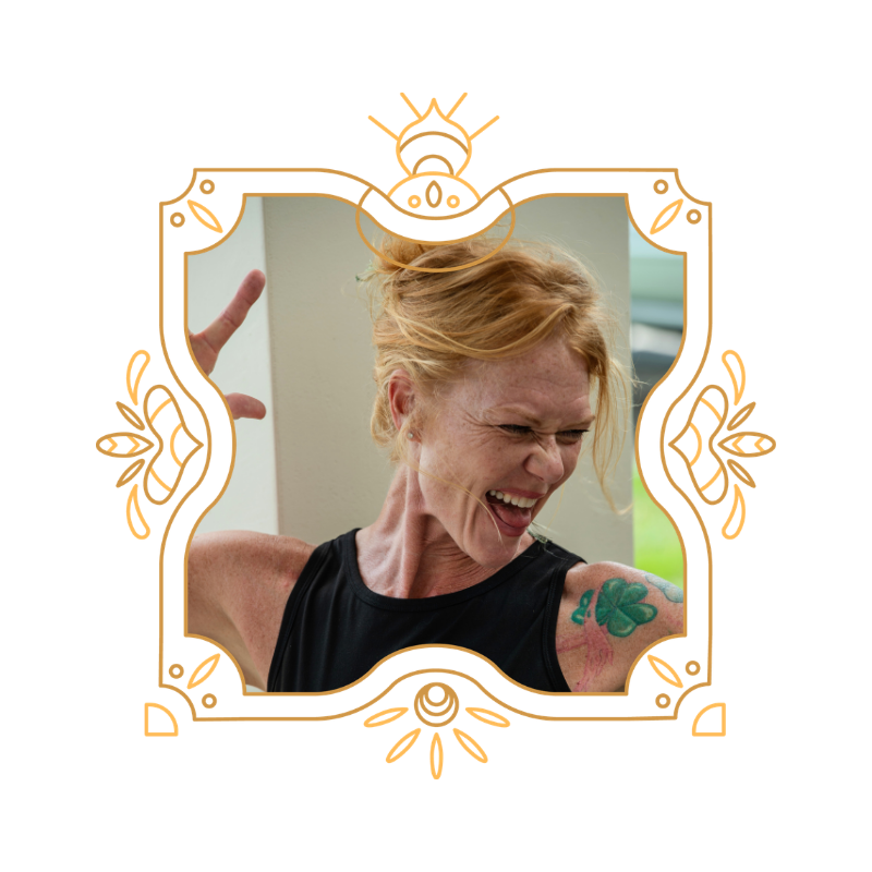 A red-haired woman with a tattoo of a four-leaf clover on her shoulder, expressing joy or excitement, making a victory sign with her hand.