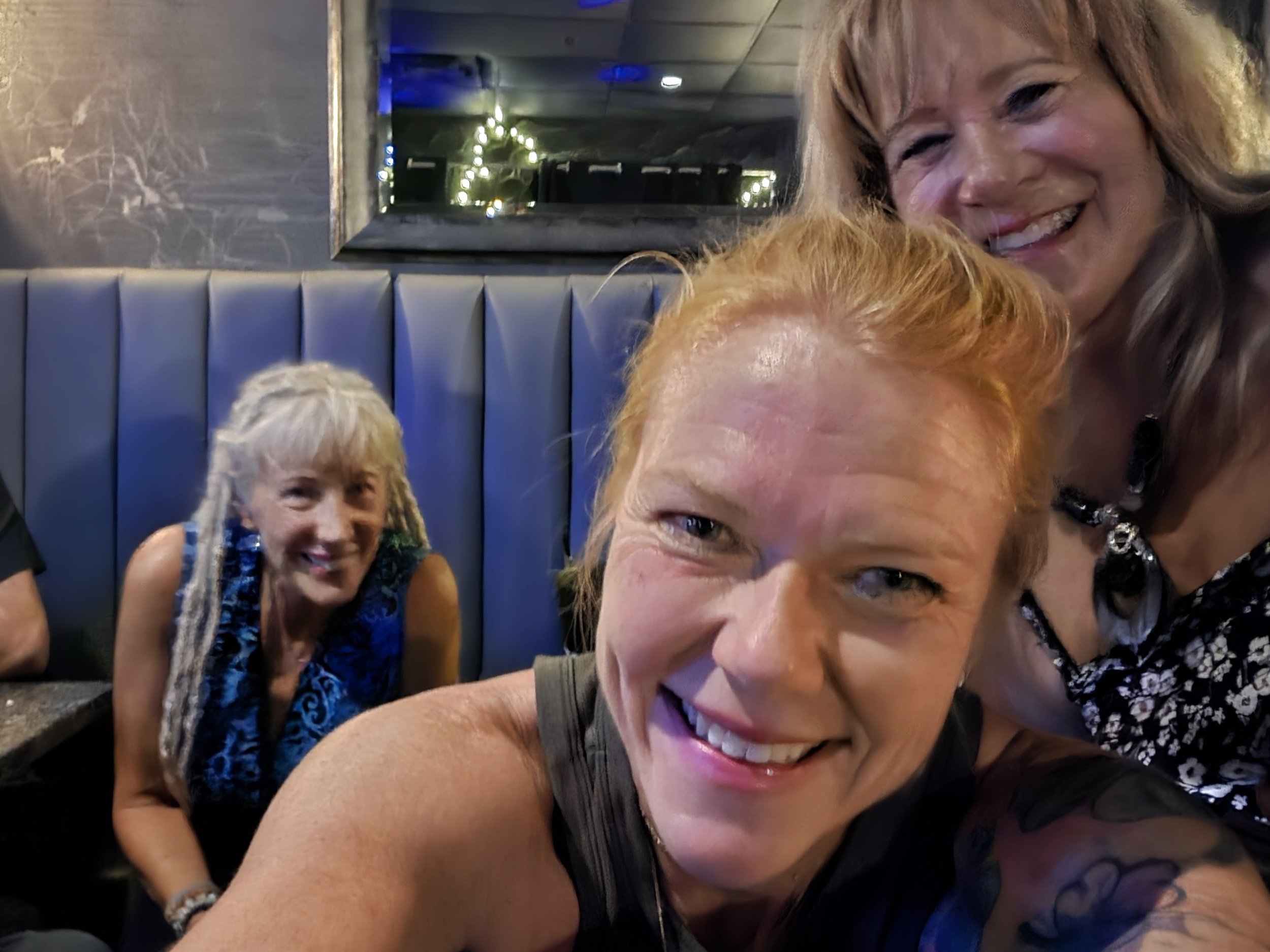 Four women smiling and taking a selfie in a dimly lit restaurant or bar with blue upholstered booth seats and decorative fairy lights in the background.