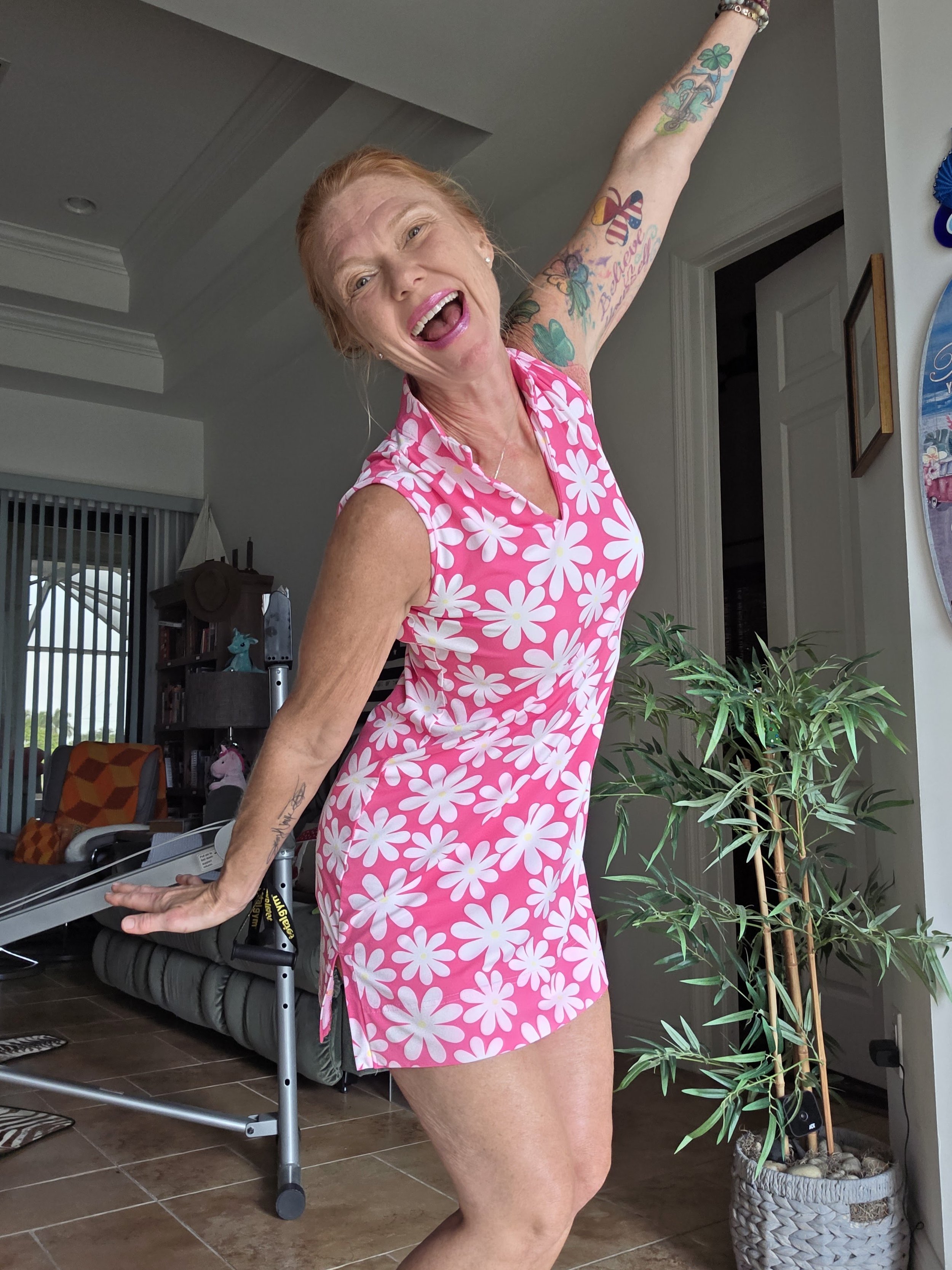 A joyful red-haired woman in a pink and white floral dress dancing in a living room.
